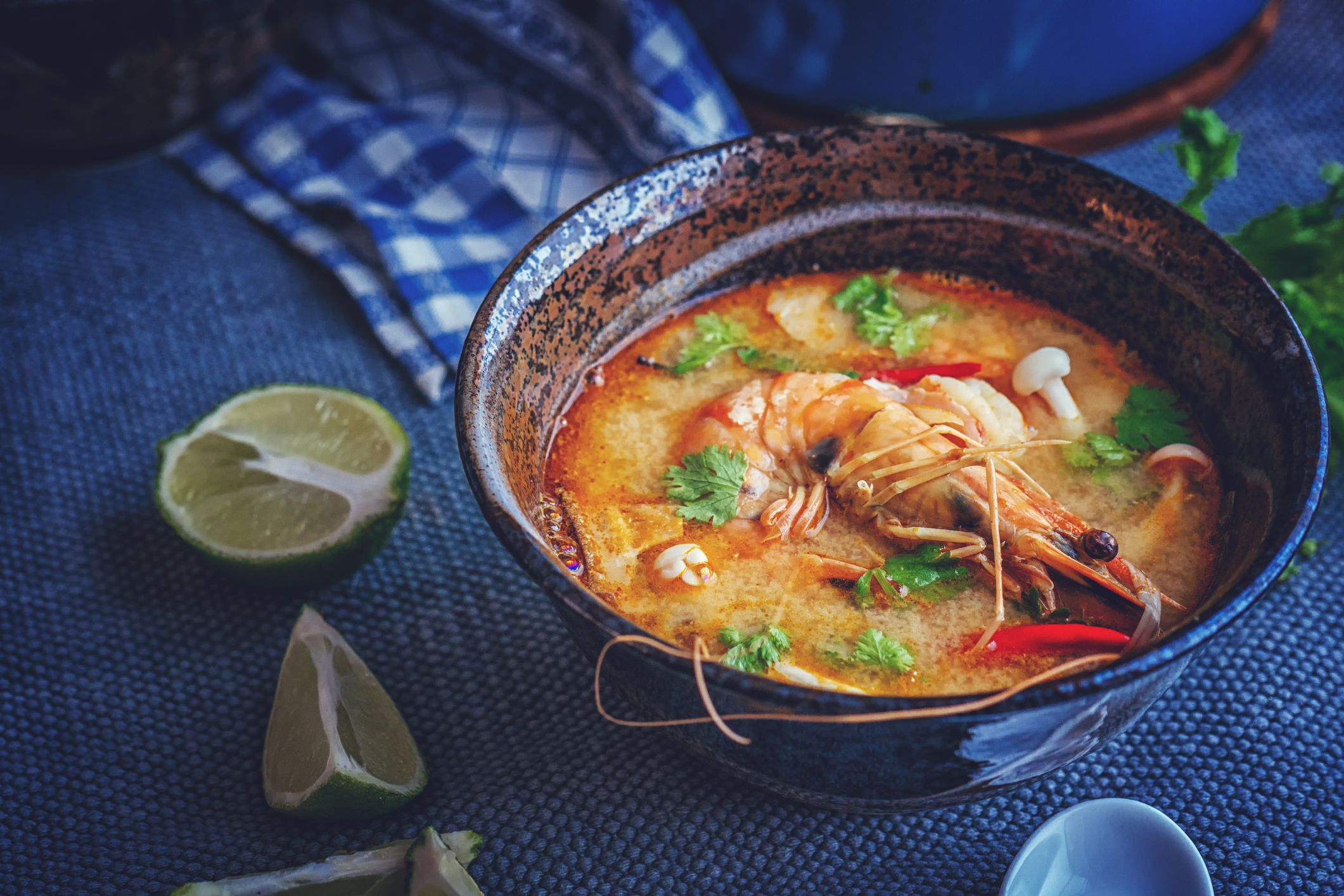 Thai coconut soup with shrimp in a vivid blue bowl on a blue table mat with a matching checkered blue napkin and lime halves