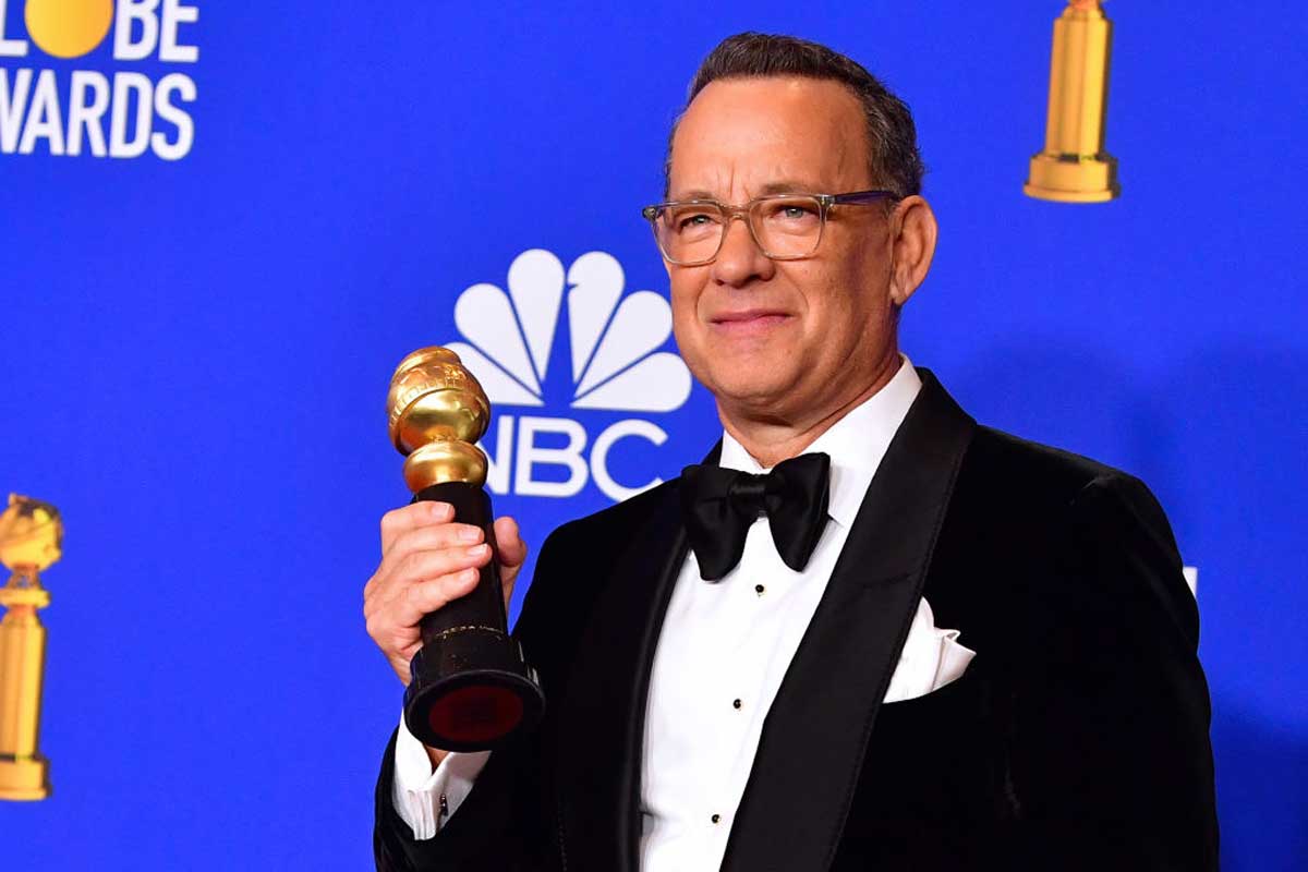 A man in a tuxedo and glasses holds a Golden Globe award and smiles in front of a blue backdrop with logos, including NBC and Golden Globe graphics, celebrating success alongside fellow celebrities.