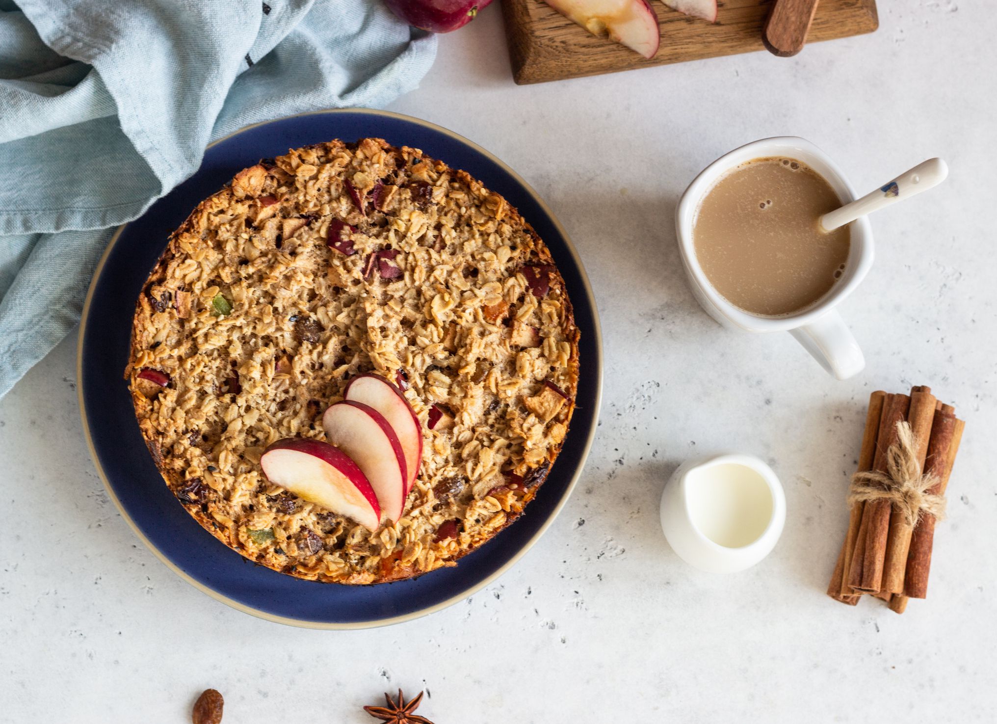 Oatmeal in a navy blue ceramic bowl with three slices of apple on the left, with a white ceramic mug of coffee and cream with a spoon in it, and a tiny white porcelain jug of milk, on a speckled white rock table, surrounded by blue napkin and slices of a