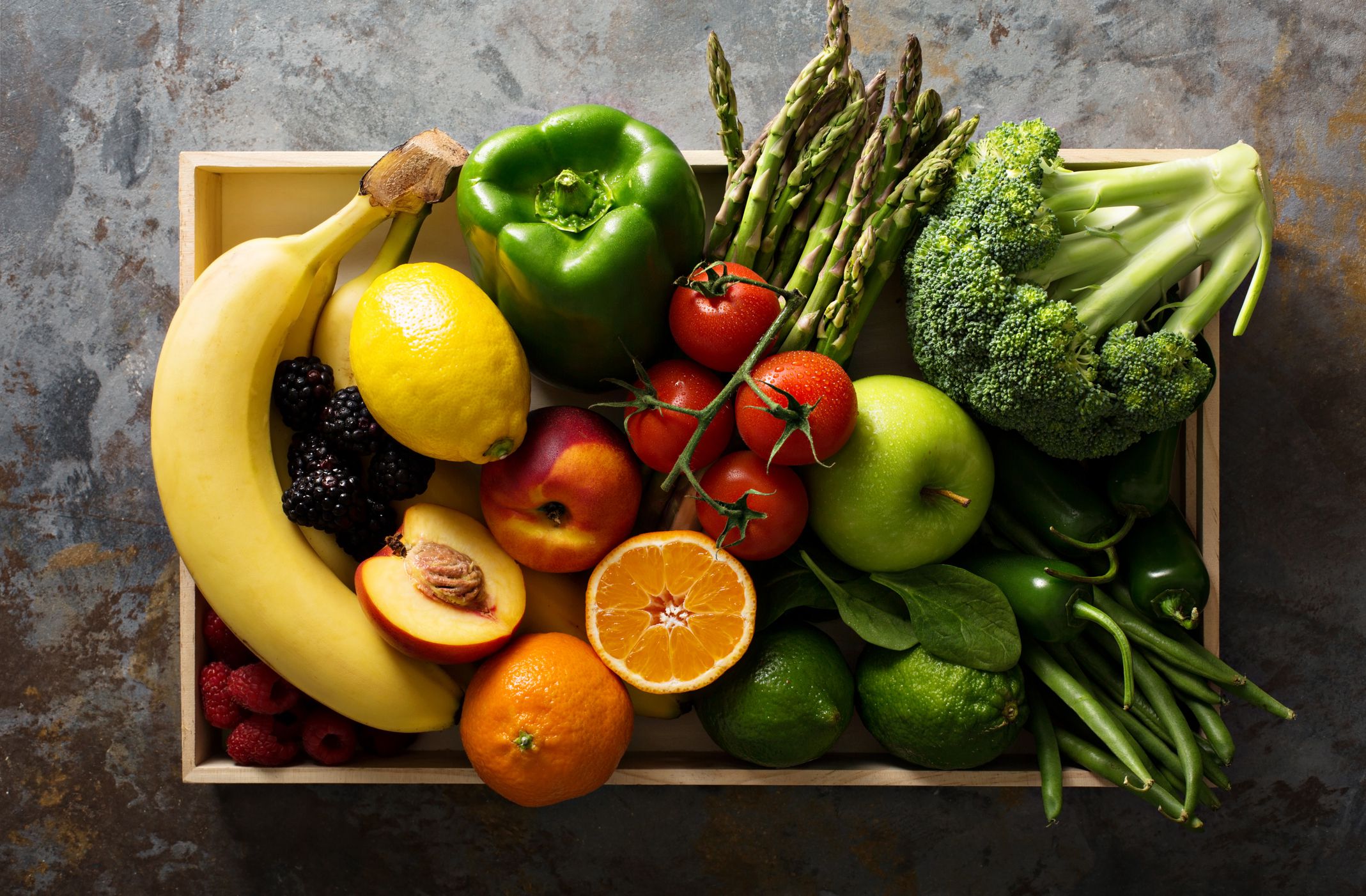 Top-view of several different kinds of fruits and vegetables in a wooden crate basket including bananas, asparagus, broccoli, oranges, and more on a rustic grey stone table