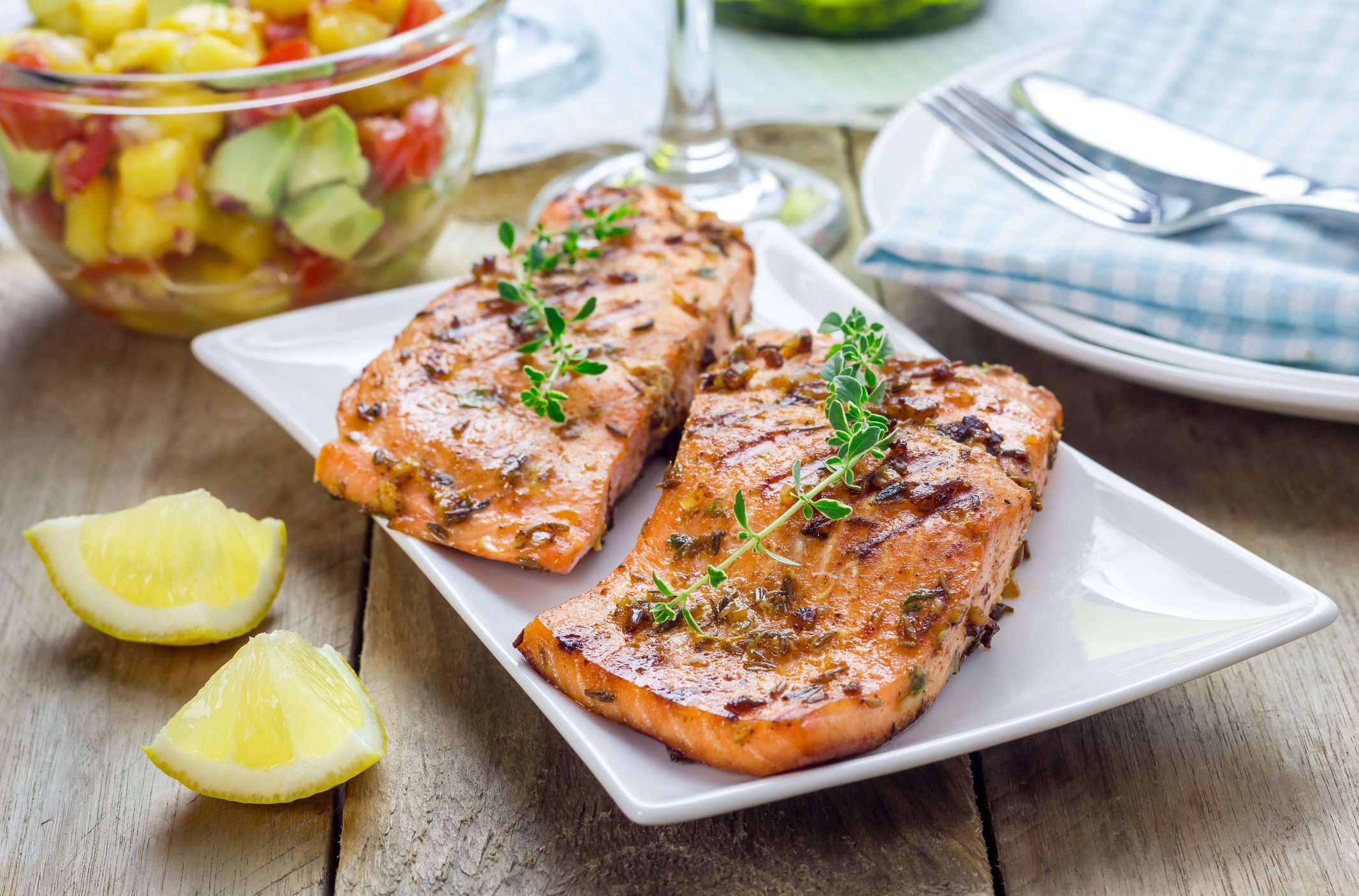 Two pieces of lemon pepper salt rub on a salmon on a white porcelain rectangle plate, surrounded by two lemon pieces, a bowl of salad, another plate with utensils and a napkin, two wine glasses on a wooden table with a blurred background