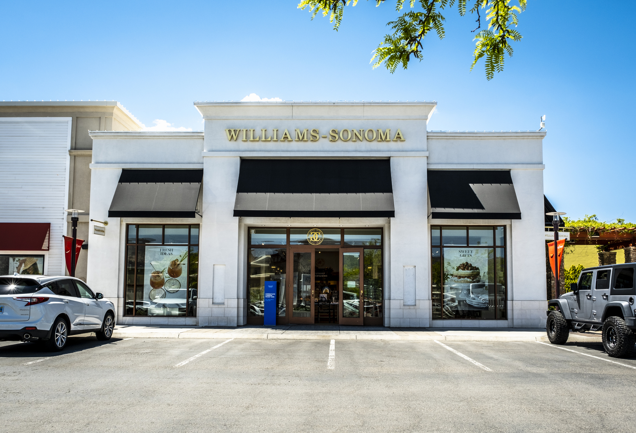 Front exterior of a Williams-Sonoma store at The Summit shopping center in South Reno, Nevada with parking spots, framed by a few tree branches on the top, on a bright sunny day