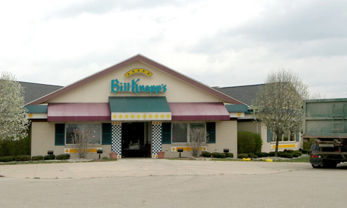 Exterior view of a Bill Knapp's, one of the classic old restaurant chains, with a peaked roof, maroon awnings, and a yellow and blue sign above the entrance. The mostly empty parking lot is bordered by trees surrounding the building.