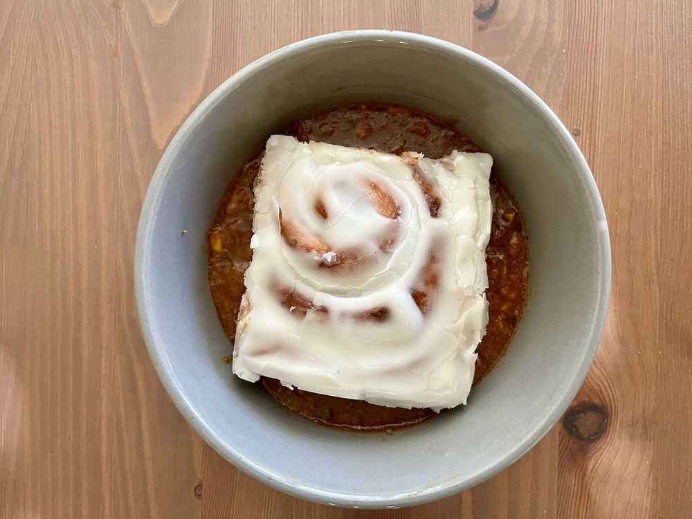 A bowl with a frosted cinnamon roll sitting on top of what appears to be chili, placed on a wooden table.