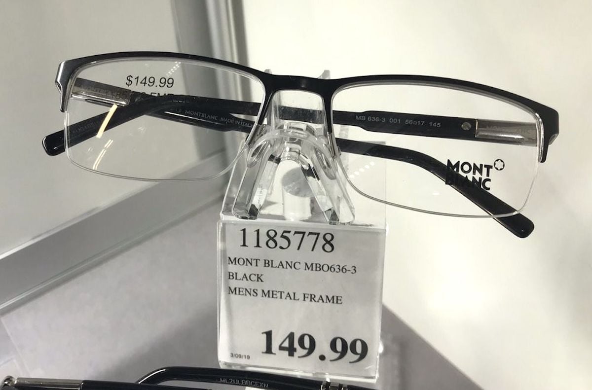 A pair of Mont Blanc men’s metal frame eyeglasses is displayed on a clear stand at Costco Optical. A price tag below shows the price as $149.99 and lists the model number, color (black), and "MEN'S METAL FRAME.