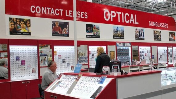 A woman stands at the counter of Costco Optical, speaking to a seated employee. Eyeglass frames are displayed in glass cases, and signs above read "Contact Lenses," "Optical," and "Sunglasses"—showcasing Costco Optical services.