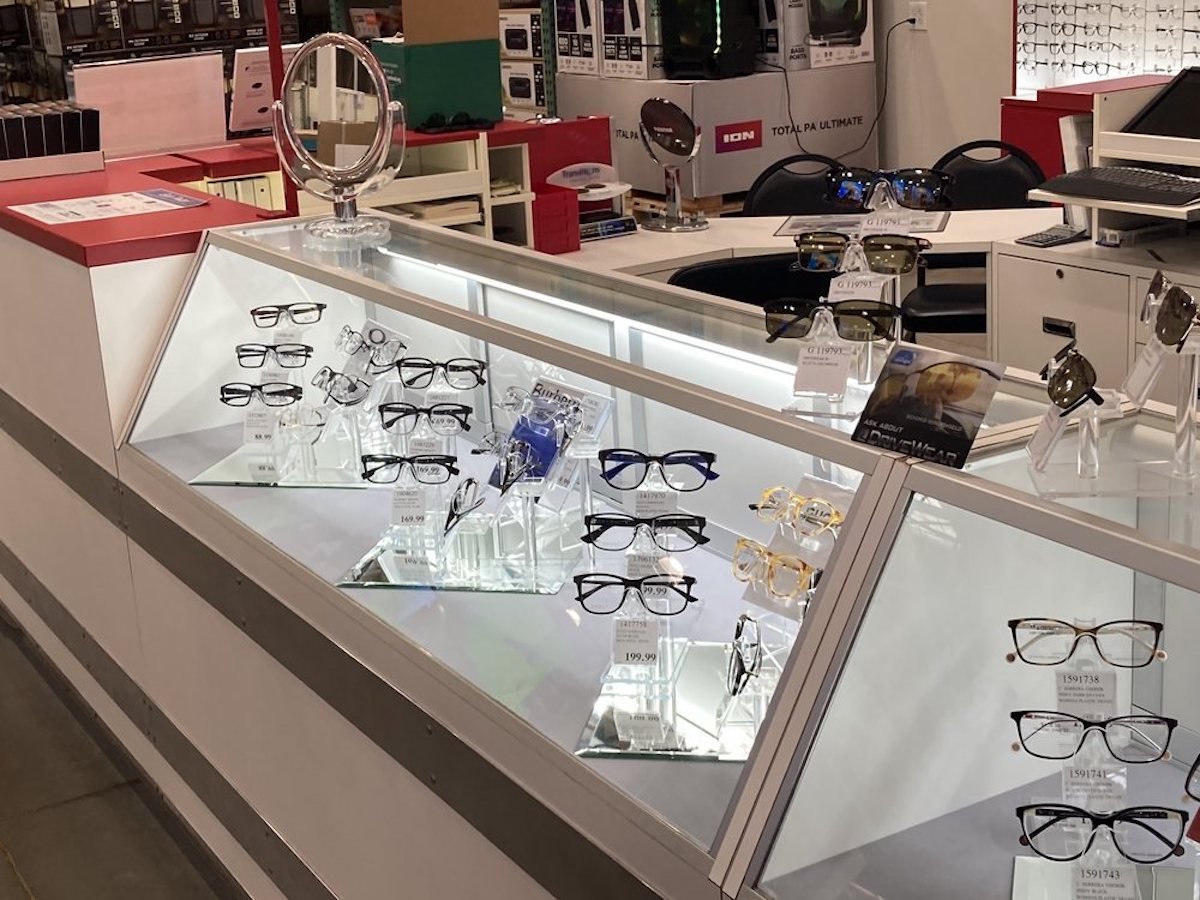 A glass display case in a Costco Optical store features eyeglasses and sunglasses on stands. The counter includes a round mirror, informational cards about Costco Optical services, and shelves with products in the background.