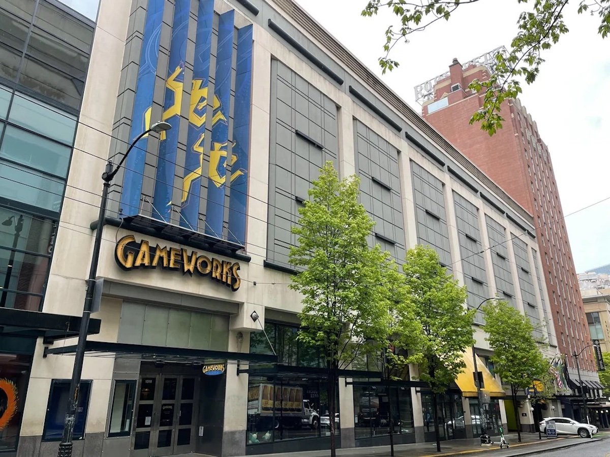 Street view of a large building with a "SEGA" vertical sign and "GameWorks" entrance, reminiscent of spots once occupied by old restaurant chains. Trees line the sidewalk, and reflections appear in the many upper-floor windows.