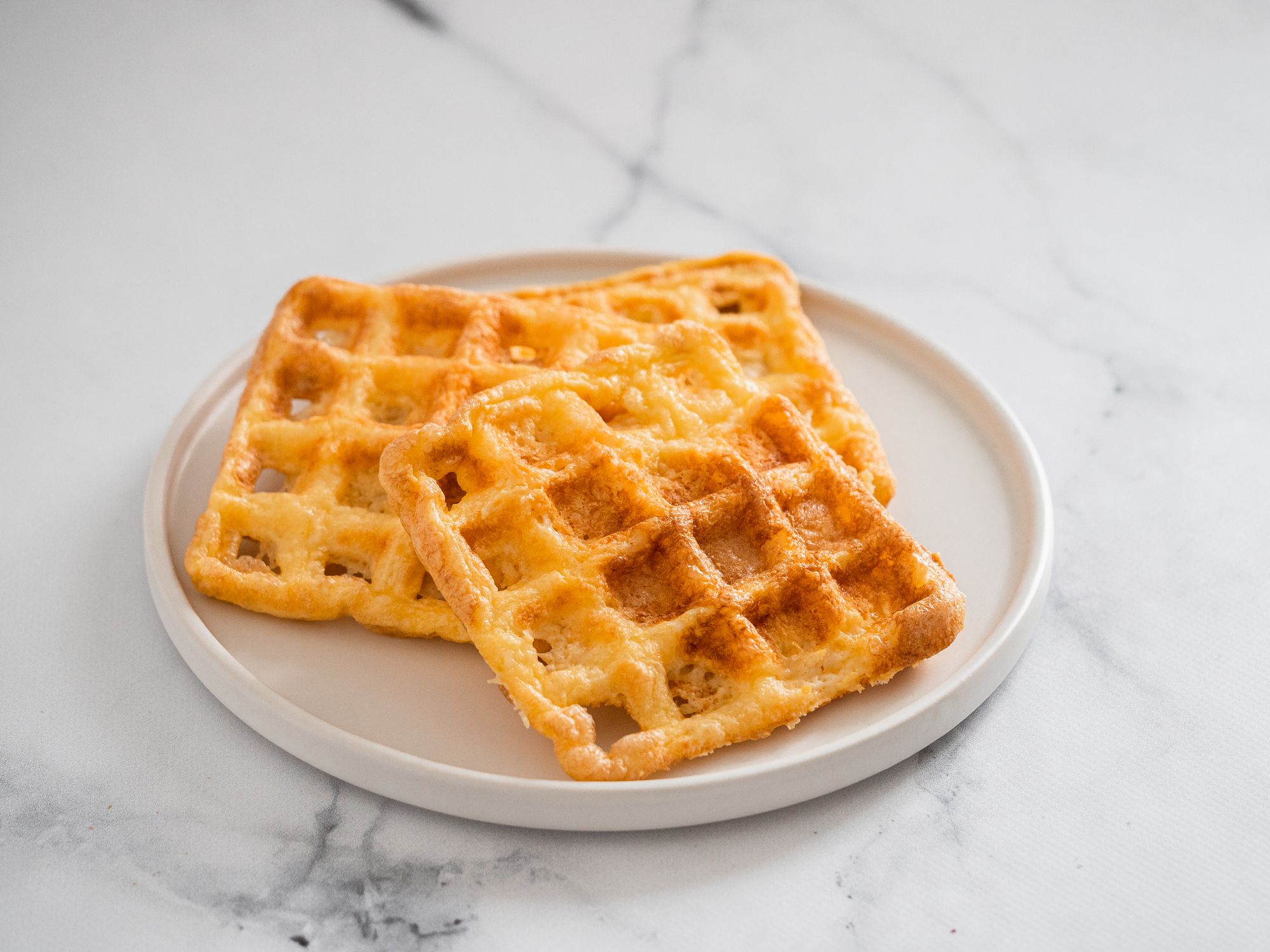 Two golden, square waffles are stacked on a round white plate, set on a white marble surface.