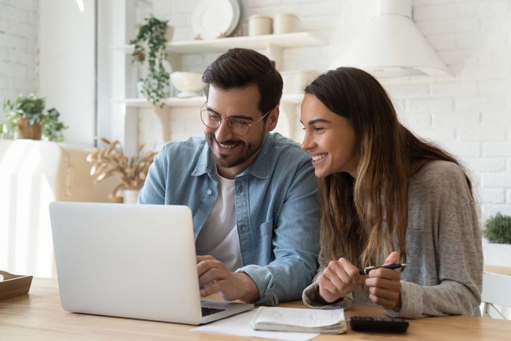 A smiling man and woman sit at a table using a laptop together. Papers, a notebook, and a pen are on the table, and they appear to be working or studying in a bright, modern kitchen.