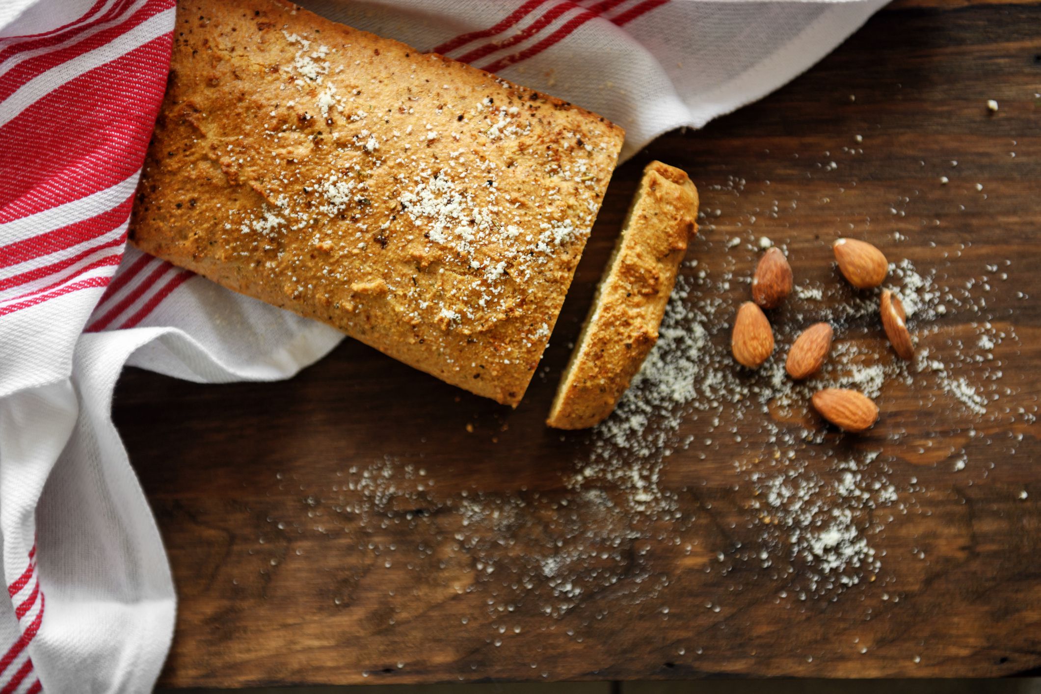 A loaf of bread partially sliced sits on a wooden surface with a red and white cloth nearby. Crumbs and several whole almonds are scattered beside the bread.