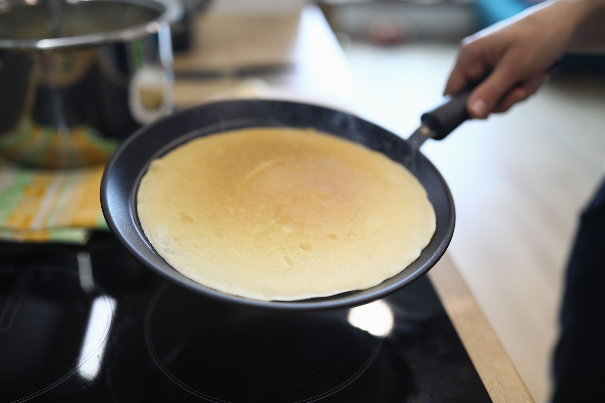 A person holds a frying pan with a freshly cooked, golden-brown crepe over a stove in a kitchen.