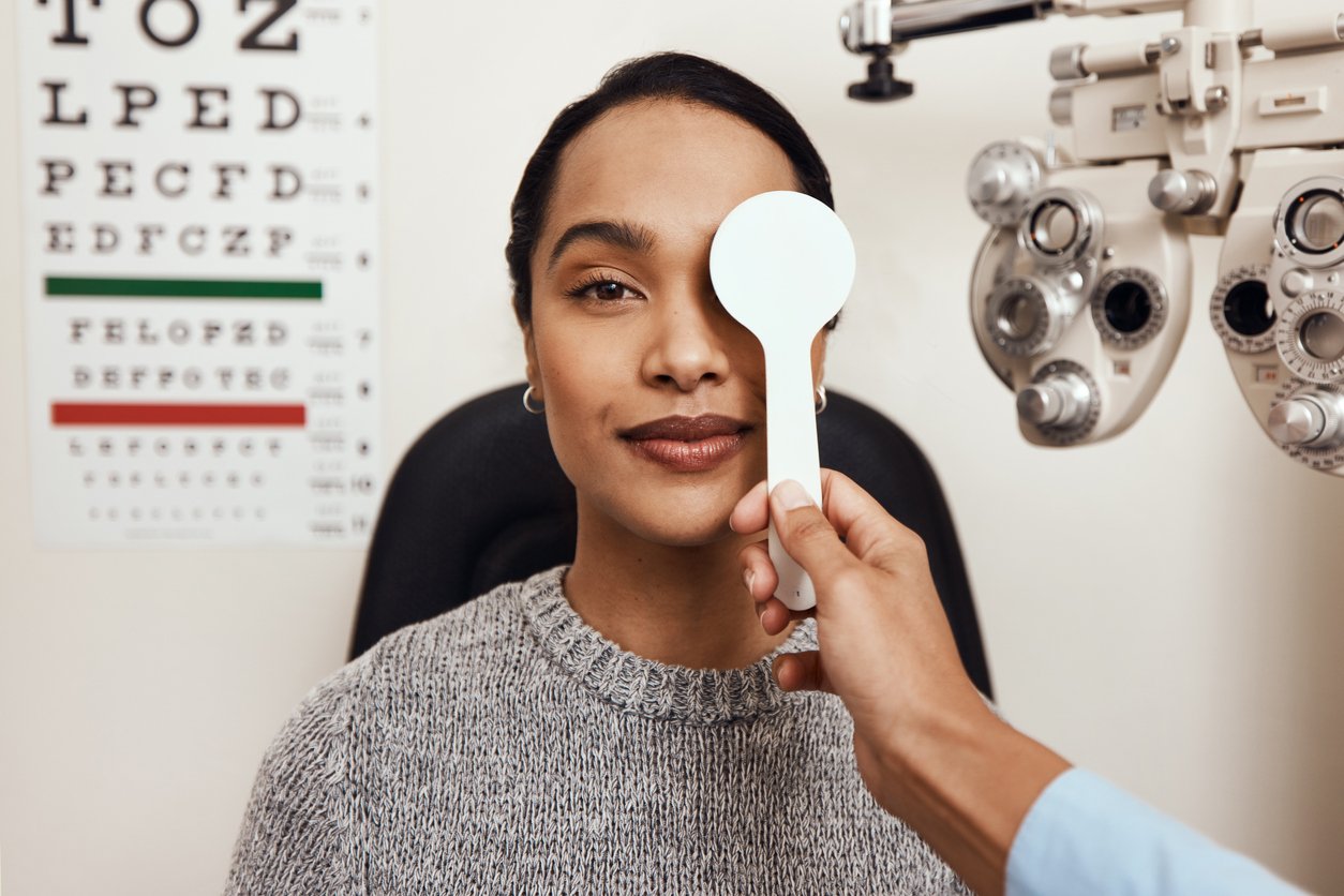 A woman sits in an eye clinic with one eye covered by an occluder, while an optometrist prepares for an eye exam. An eye chart and examination equipment are visible, showcasing Costco Optical services in the background.