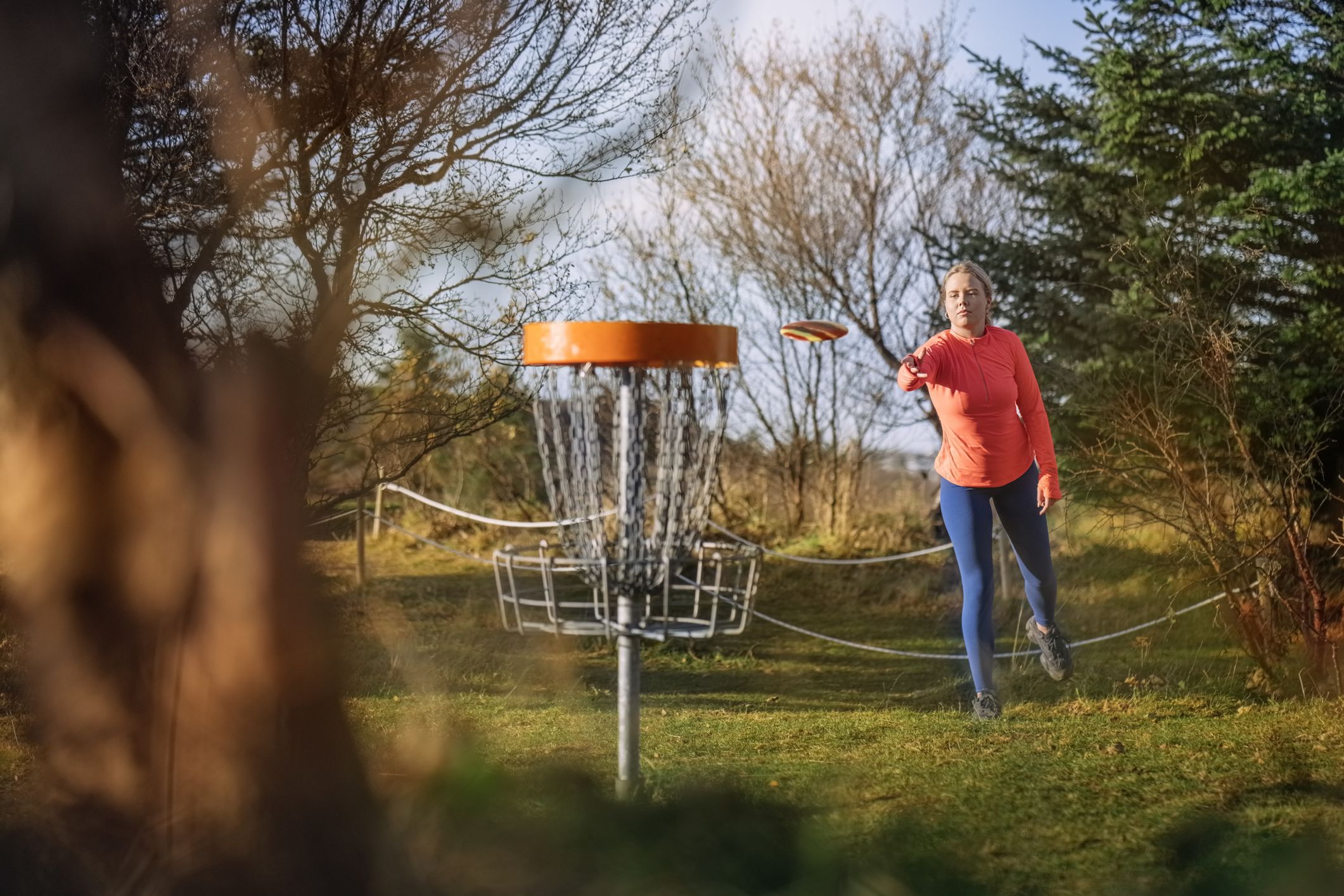 Woman tossing a disc into the basket goal. Caucasian woman playing disc golf game outdoors in a park.