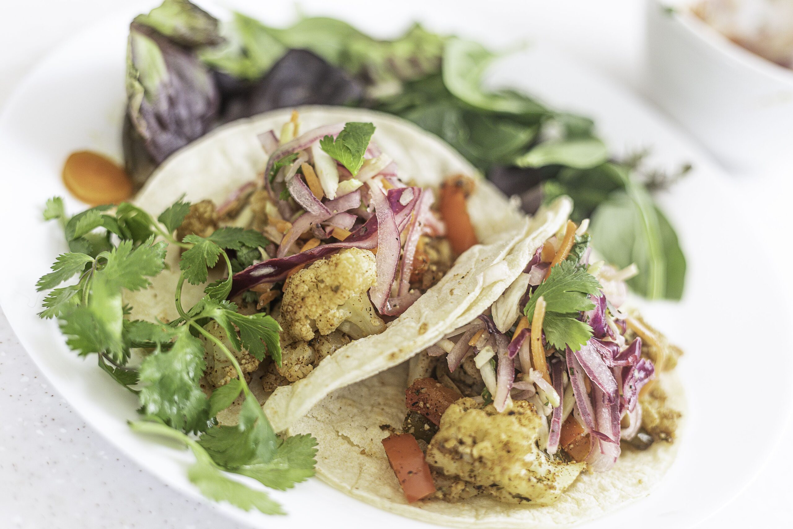 Two soft tacos filled with seasoned cauliflower, pickled onions, slaw, and fresh cilantro, served on a white plate with a side of mixed greens.