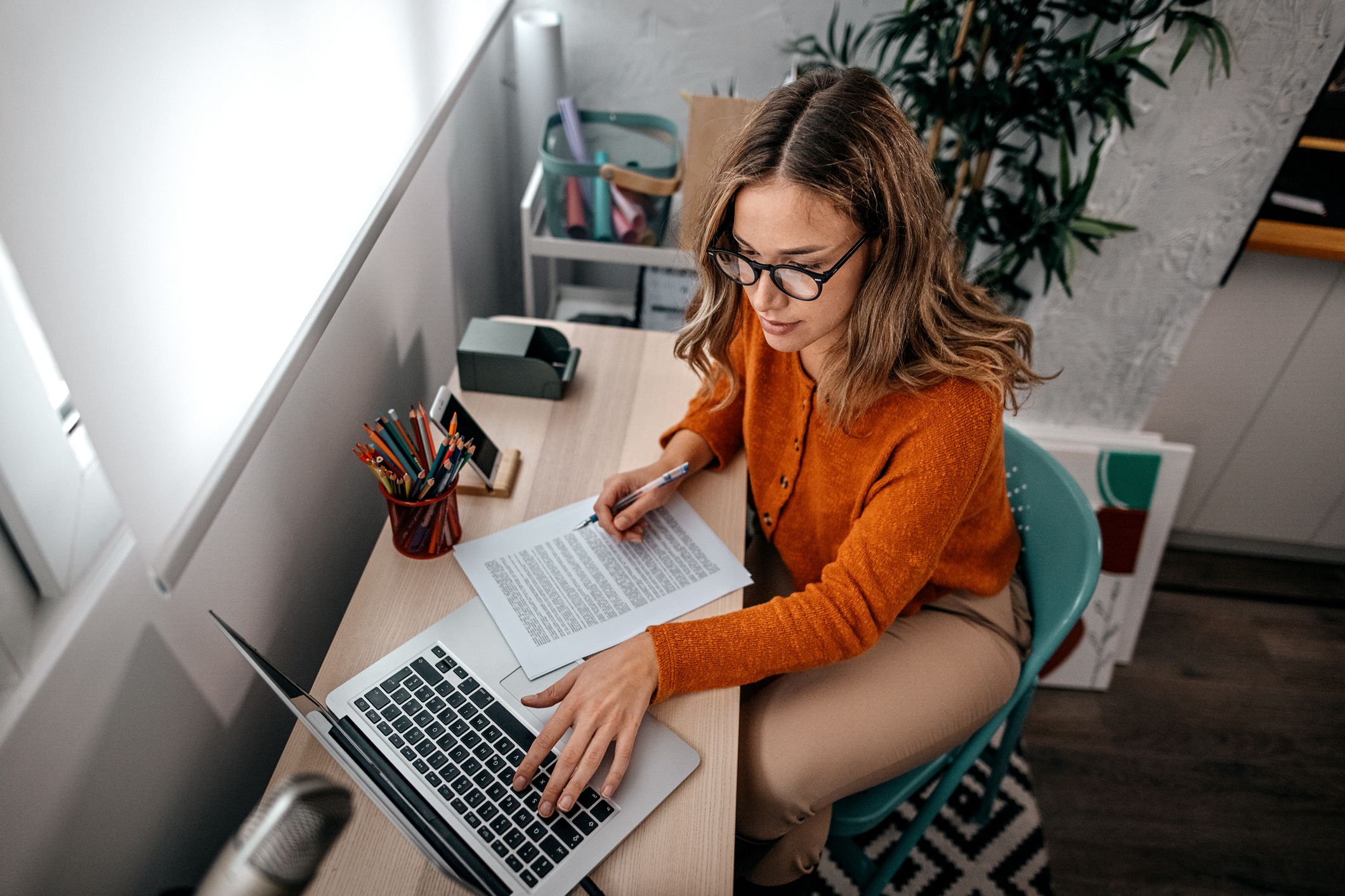 Young woman with spectacles working on laptop while reading with notes at home while podcasting