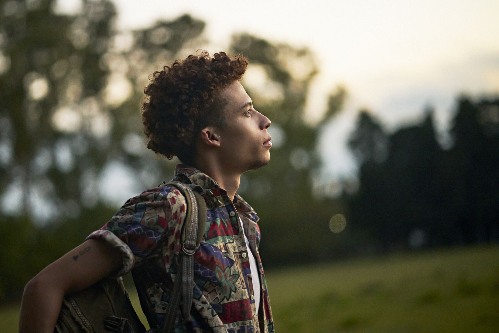 Waist-up profile view of man with curly brown hair wearing casual clothing and backpack looking away from camera with contented expression of hope.