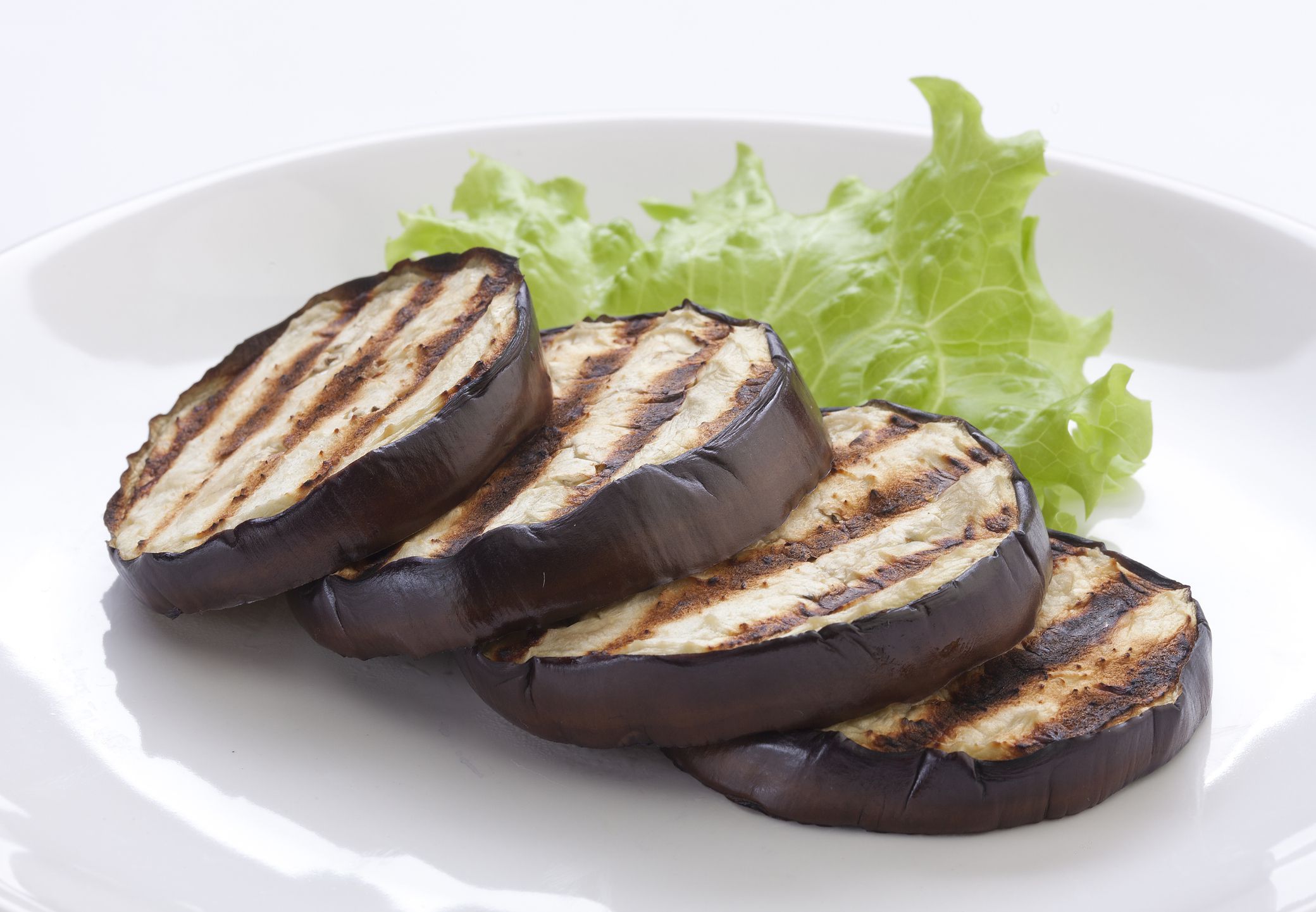 Four slices of grilled eggplant with visible grill marks are arranged in a row on a white plate, with a piece of green leaf lettuce in the background for garnish.