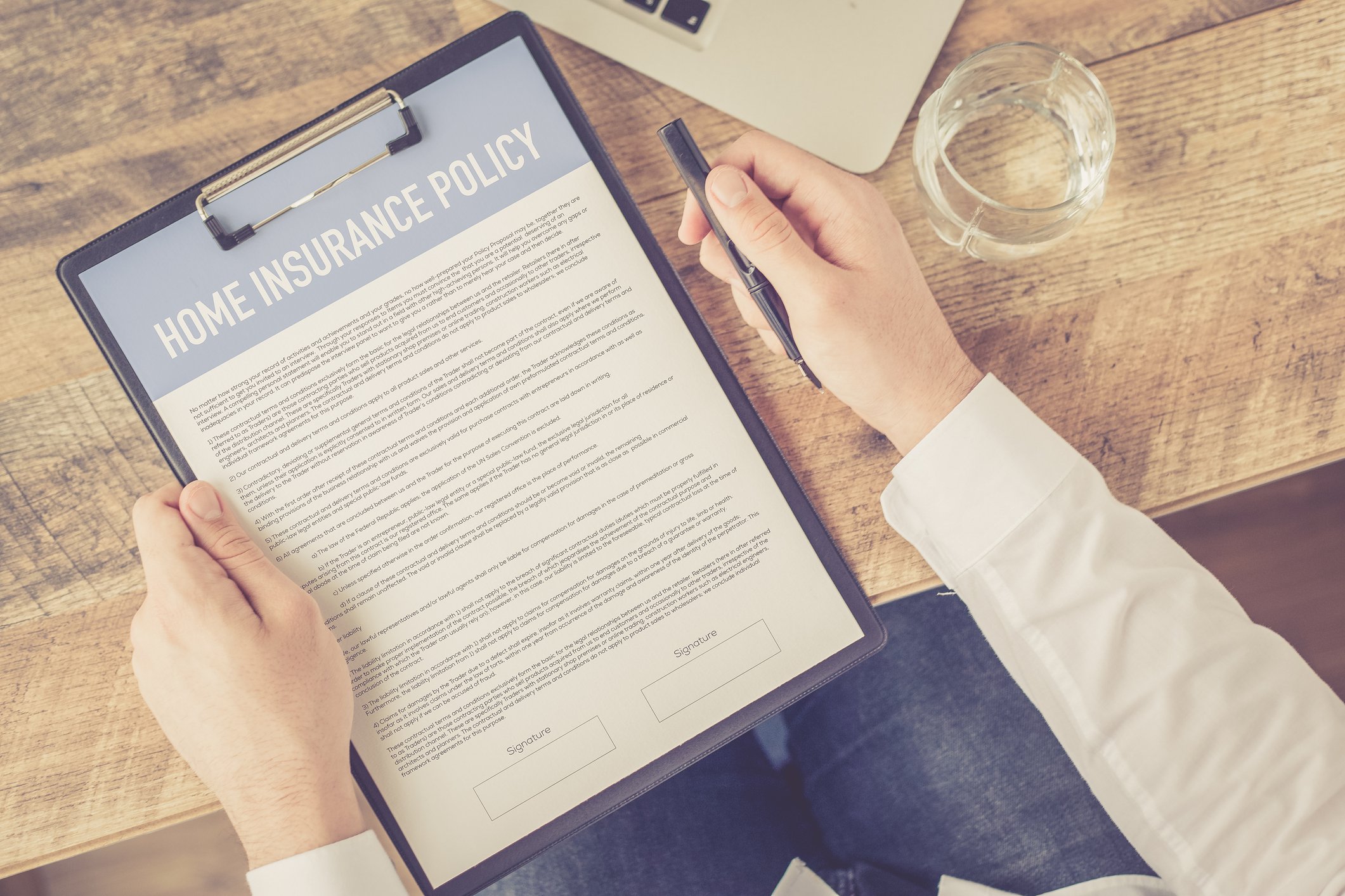 A person holding a clipboard with a home insurance policy document, a pen, and preparing to sign it at a wooden desk with a glass of water and part of a laptop visible.
