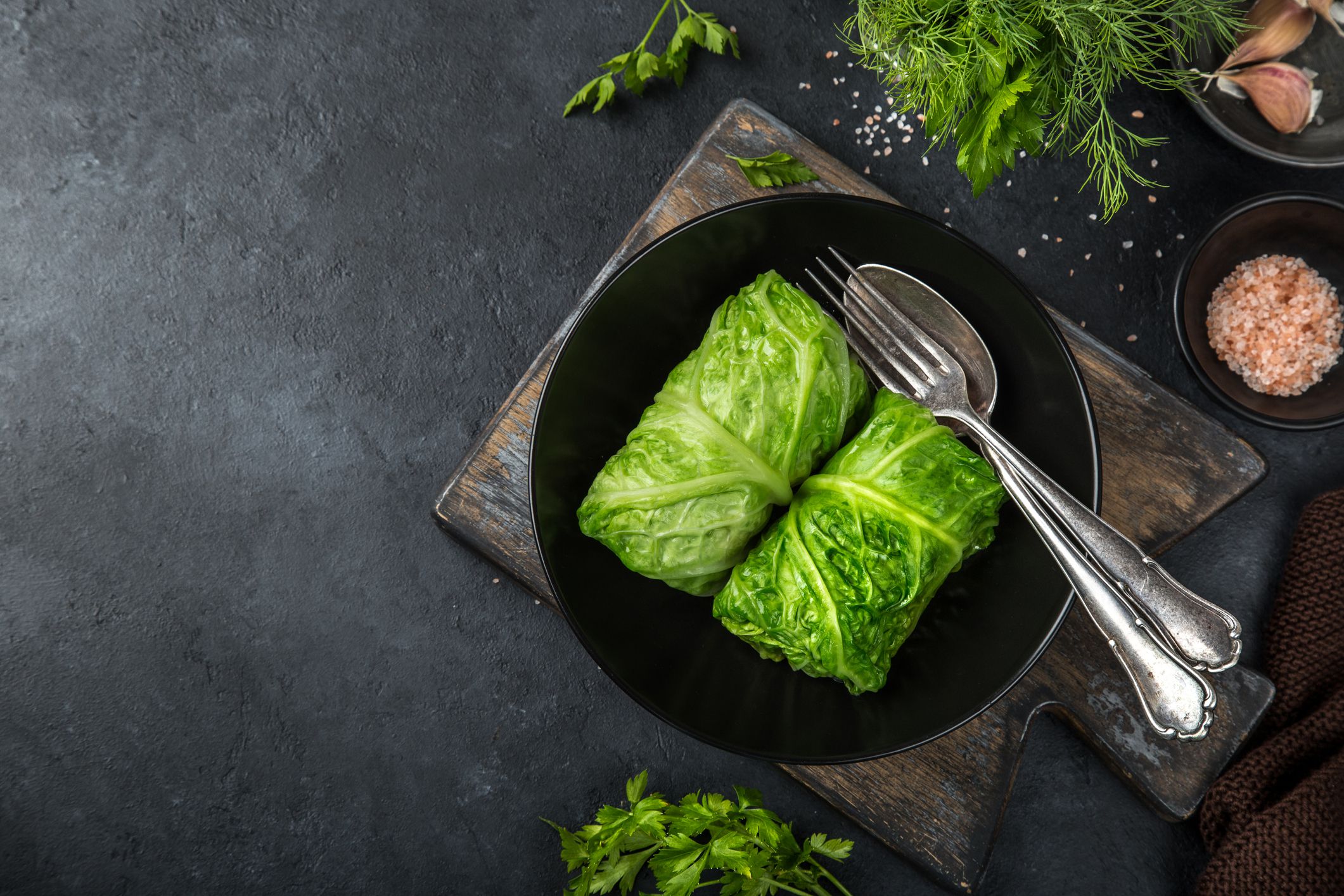 A black bowl with two cabbage rolls, topped with a fork and spoon, sits on a wooden board. Fresh herbs and a small bowl of pink salt are arranged around it on a dark background.