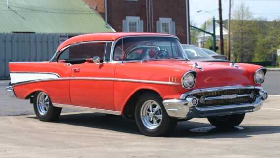 A bright red classic 1957 Chevrolet Bel Air coupe with chrome trim and whitewall tires is parked on a sunny day in front of a brick building—an iconic contrast to the average car price by year seen in today’s market.