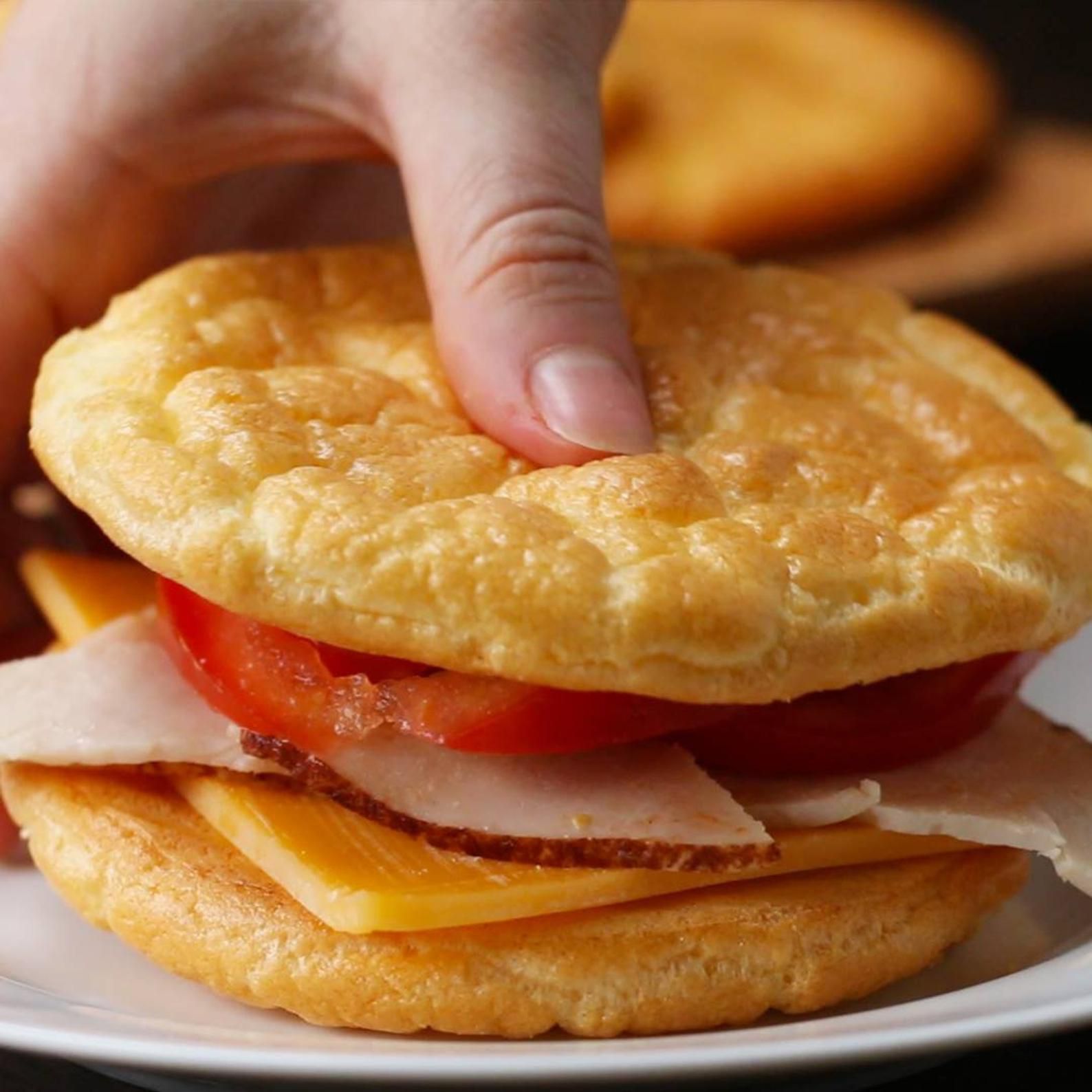 A hand holding a sandwich with fluffy, golden bread, filled with slices of cheddar cheese, turkey, and tomato on a white plate.