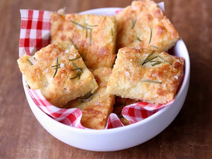 A white bowl lined with red and white checkered cloth holds several square pieces of golden-brown focaccia bread, topped with rosemary and sea salt, on a wooden surface.