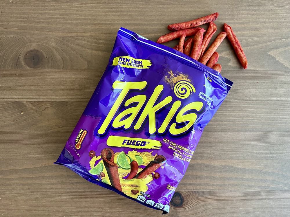 A blue and purple bag of Takis Fuego snacks lies on a wooden surface, partially open with several rolled spicy chips spilling out. The packaging shows images of the snacks and lime slices.