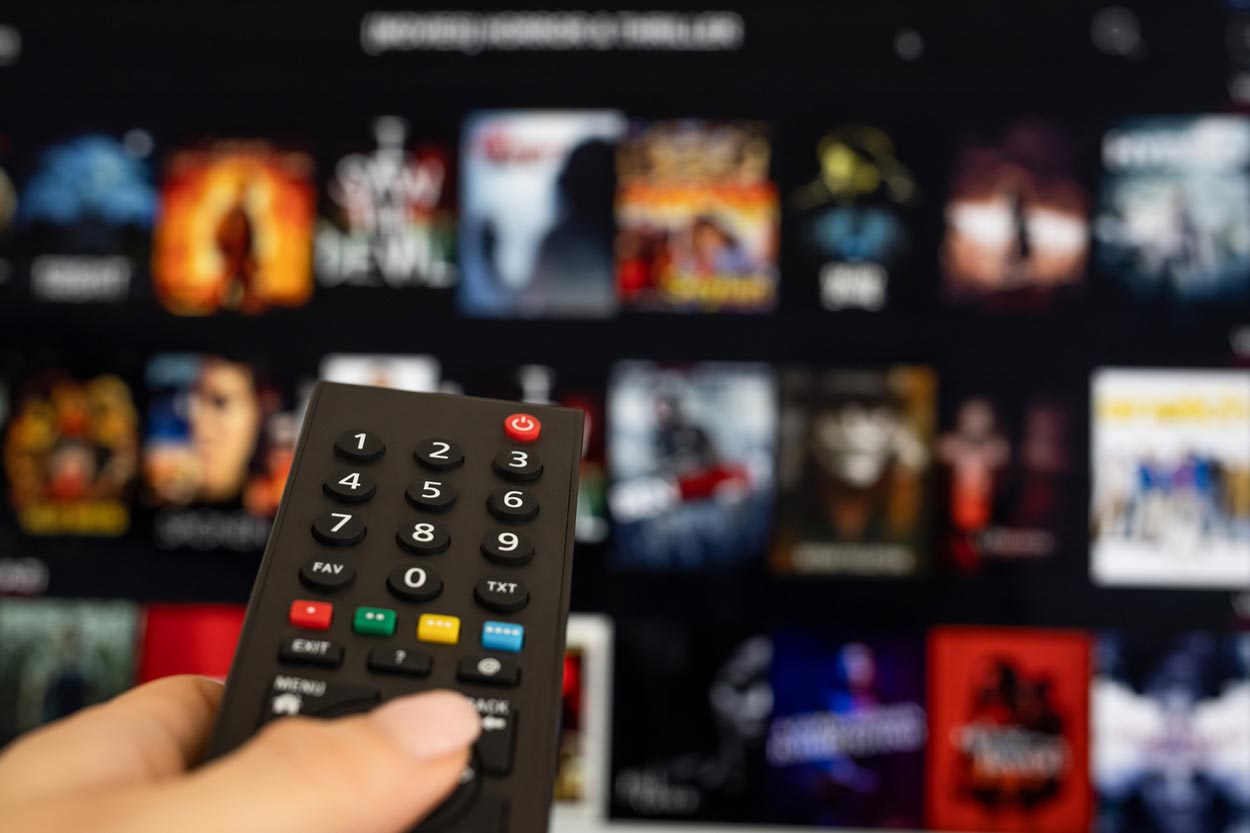 A hand holds a TV remote control pointed at a blurred television screen displaying a streaming service menu with various movie and TV show thumbnails.
