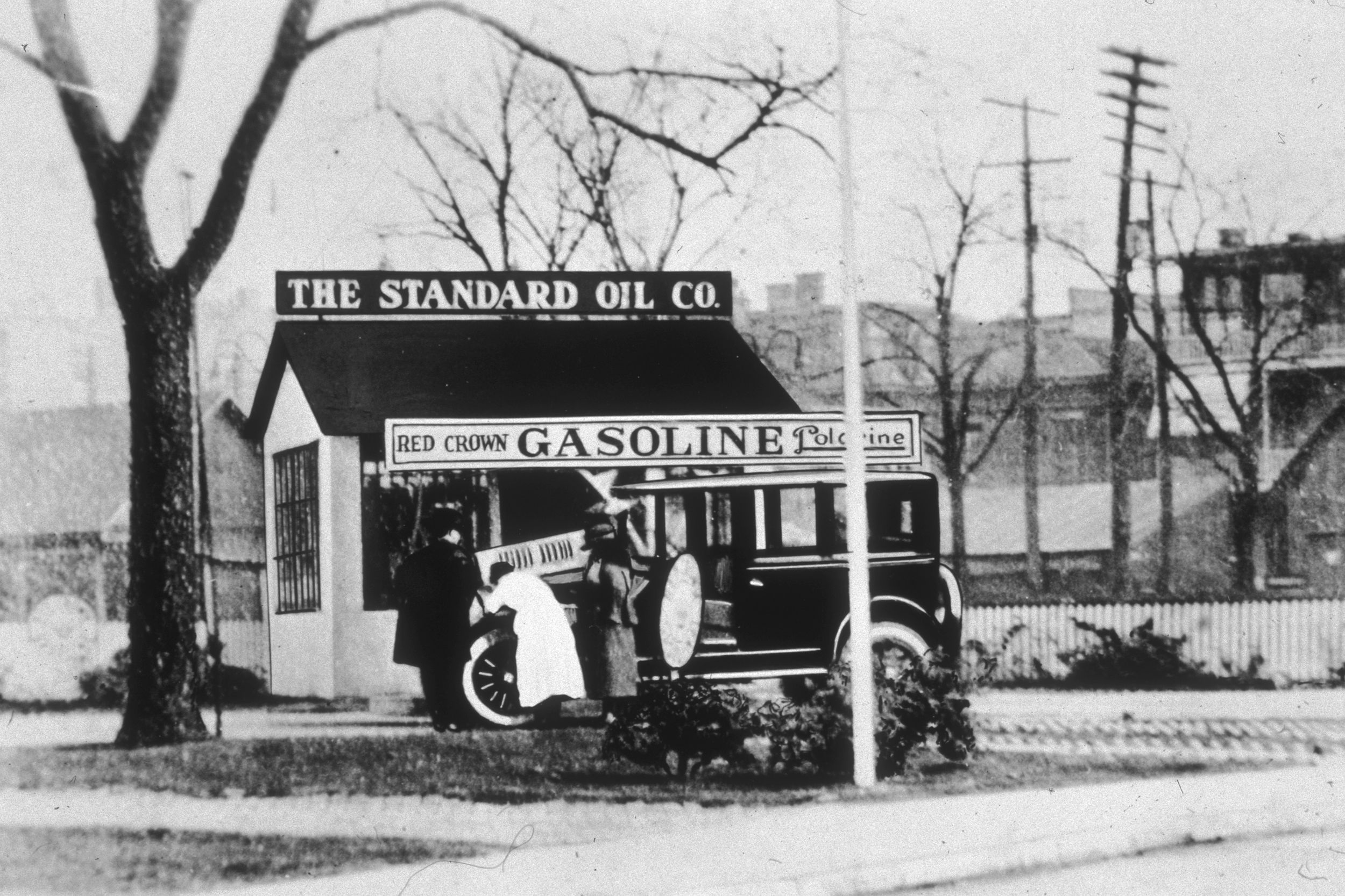 Three people look under the hood of an automobile in front of a Standard Oil gas station.