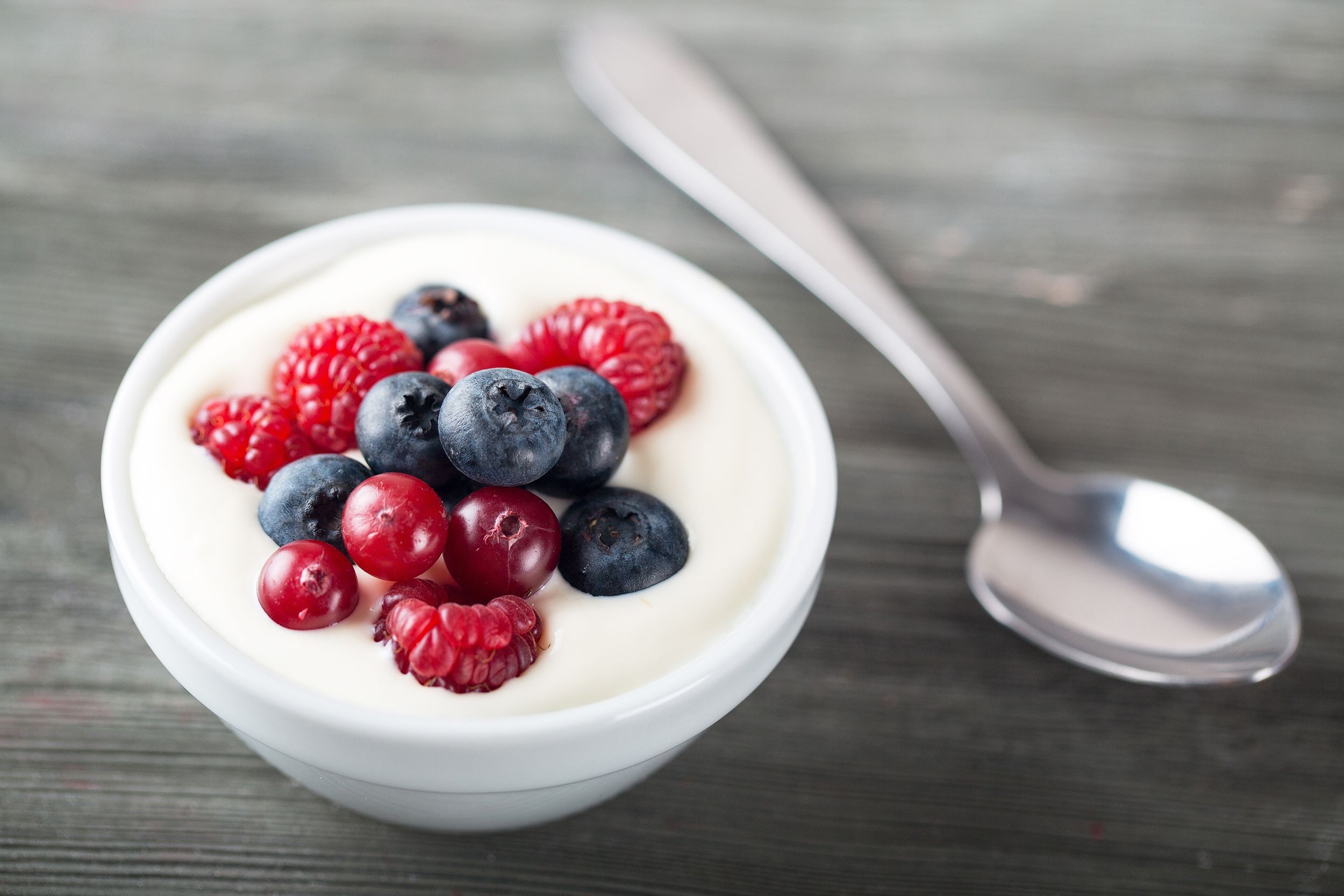 A small glass bowl of fruit & yogurt with a spoon on a grey wooden table