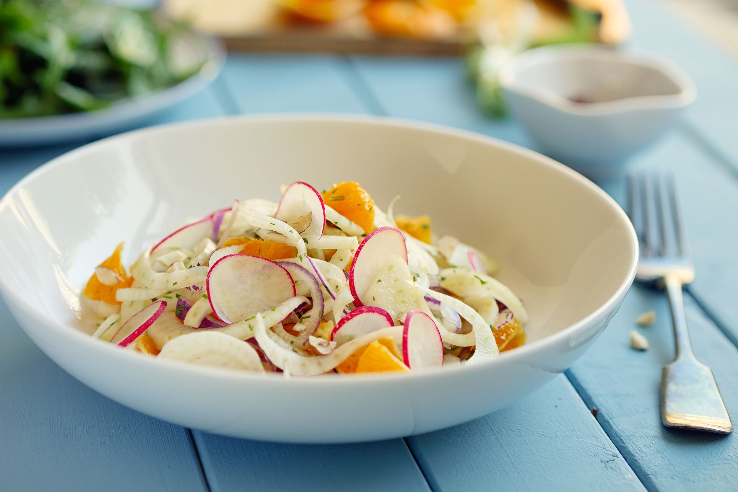 Celery, onion & orange salad in a white bowl with a fork on the right side on a light blue wooden table with a blurred background of a salad, conditments, and food