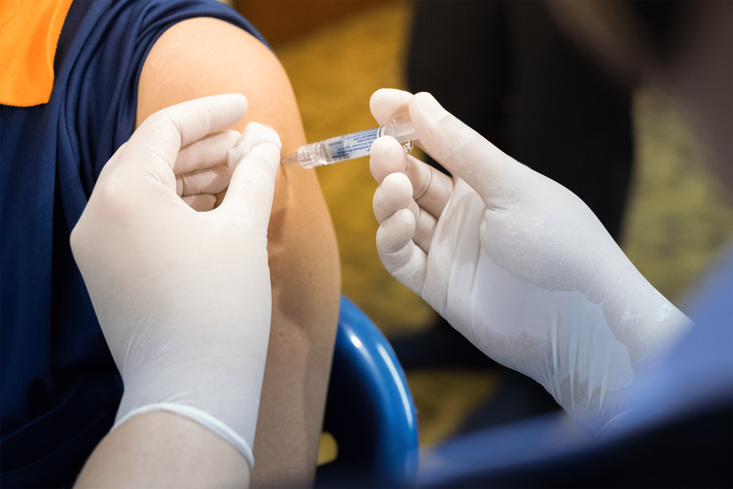 Closeup of gloved hands of a nurse giving a patient’s arm a flu shot, selective focus, patient is sitting in a blue chair, ground is blurred