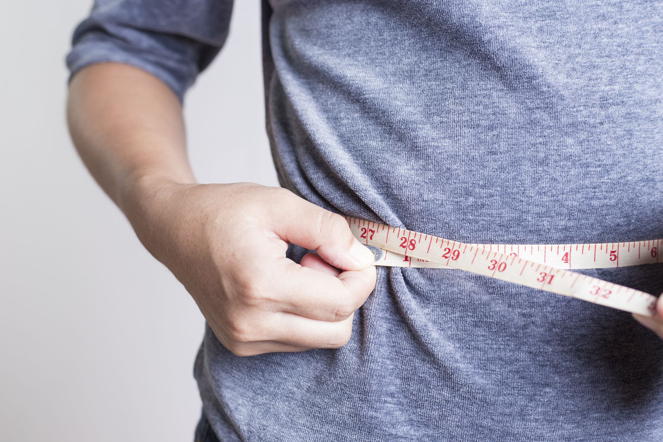 Closeup of woman in a grey shirt measuring her waist with a measuring tape in inches on a light grey background