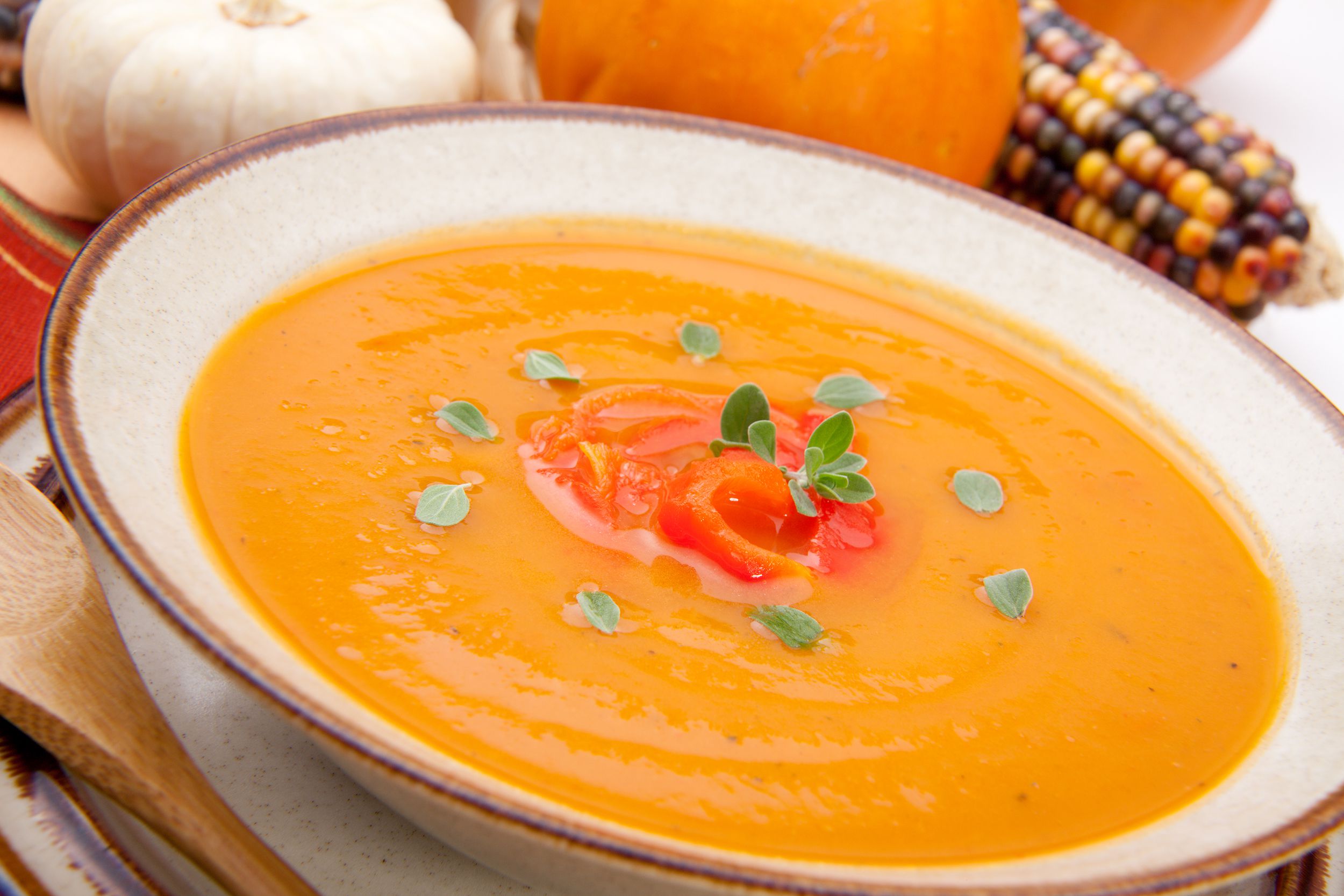 Closeup of butternut & red pepper soup garnished with fresh oregano in a ceramic bowl with fall vegetables pumpkin and corn in the background