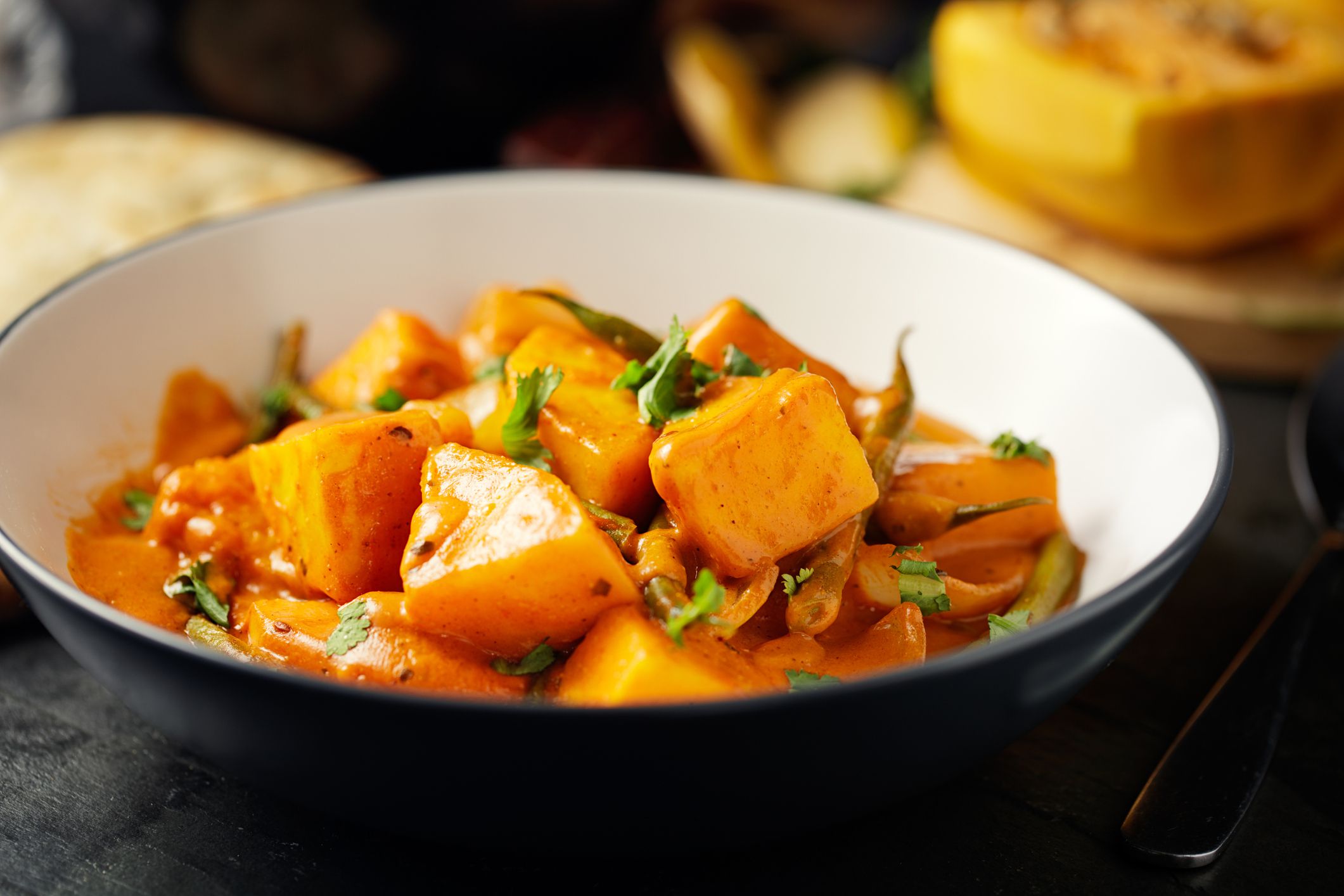 Closeup of curried winter squash in white and black bowl with a blurred background of ingredients and food