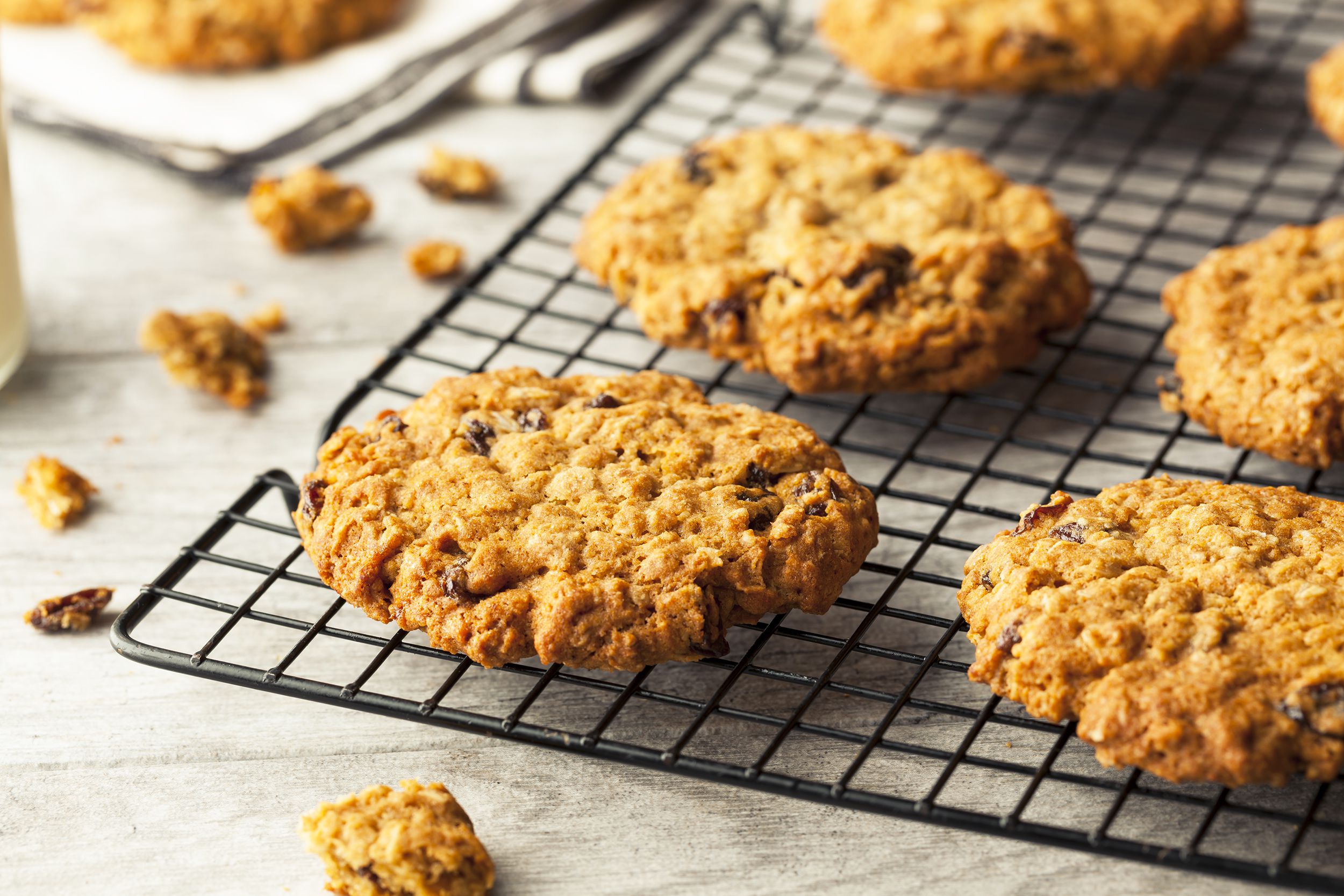 Closeup of six oatmeal raisin cookies on a black baking grid with cookie crumbs on a natural wooden table with a blurred background of cookies and napkins