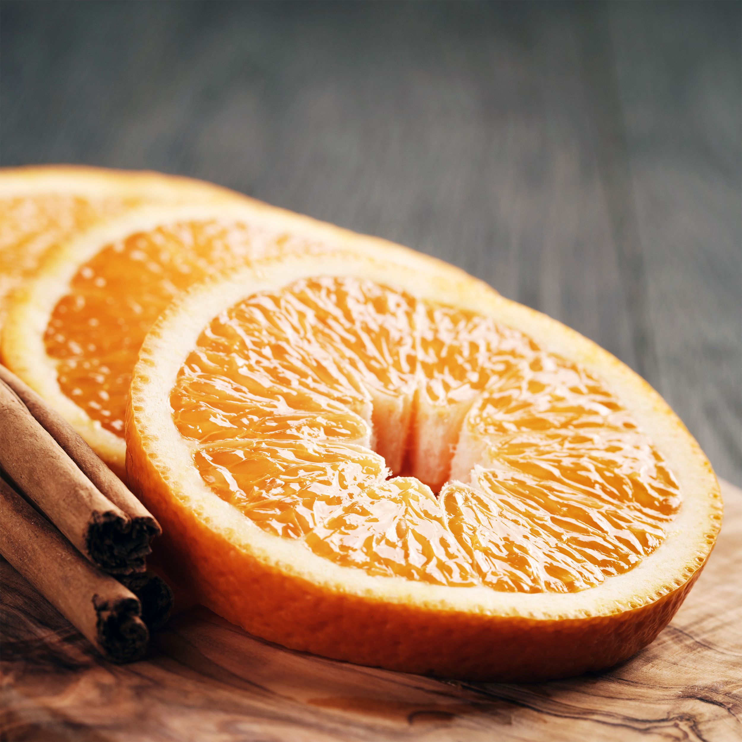 Closeup of three slices of cinnamon oranges with sticks of cinnamon on a wooden cutting board with a blurred background of a wooden grey table