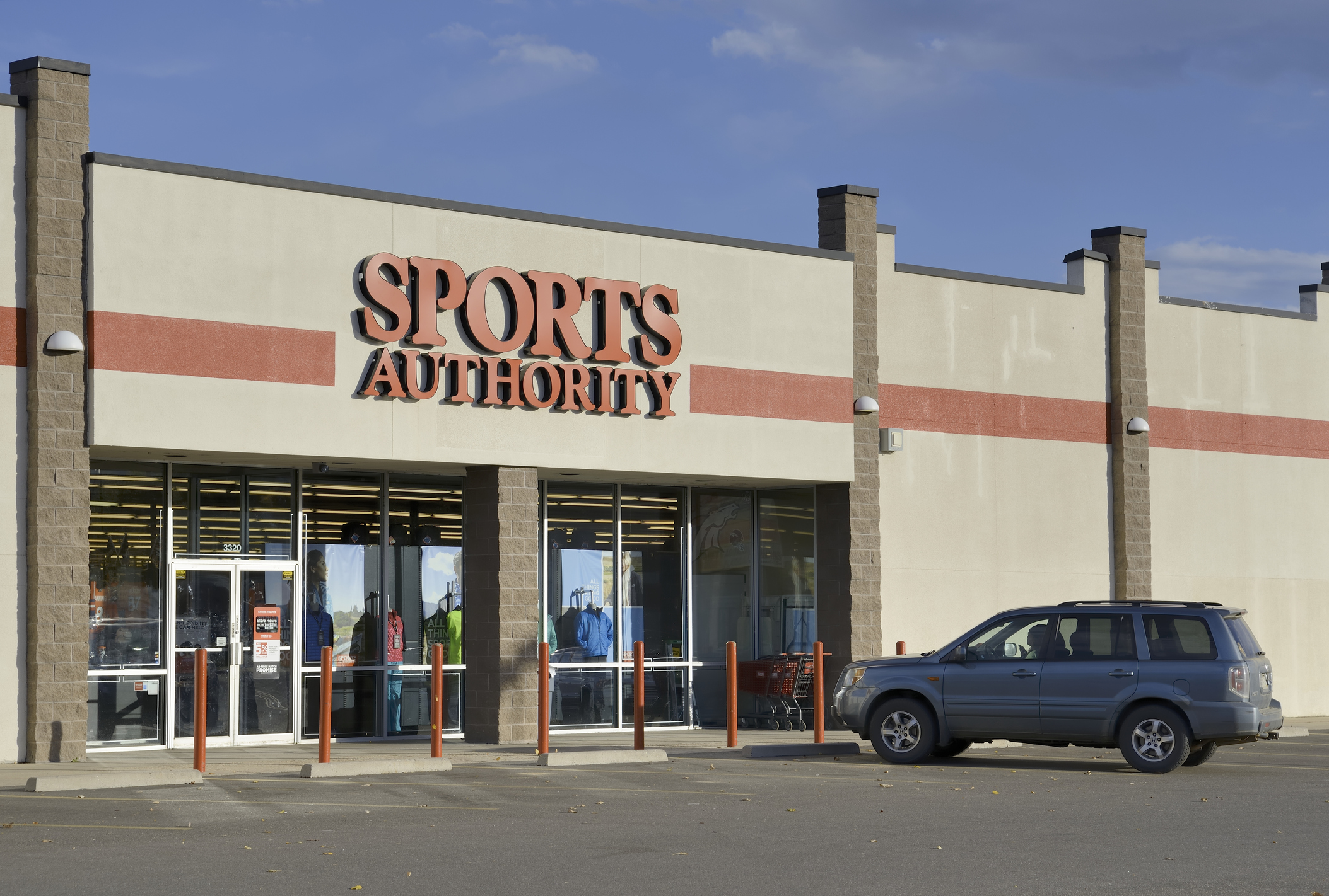 Front exterior of Sports Authority, Boulder, Colorado with one car parked in parking spots that line the sidewalk, with a blue sky in the background