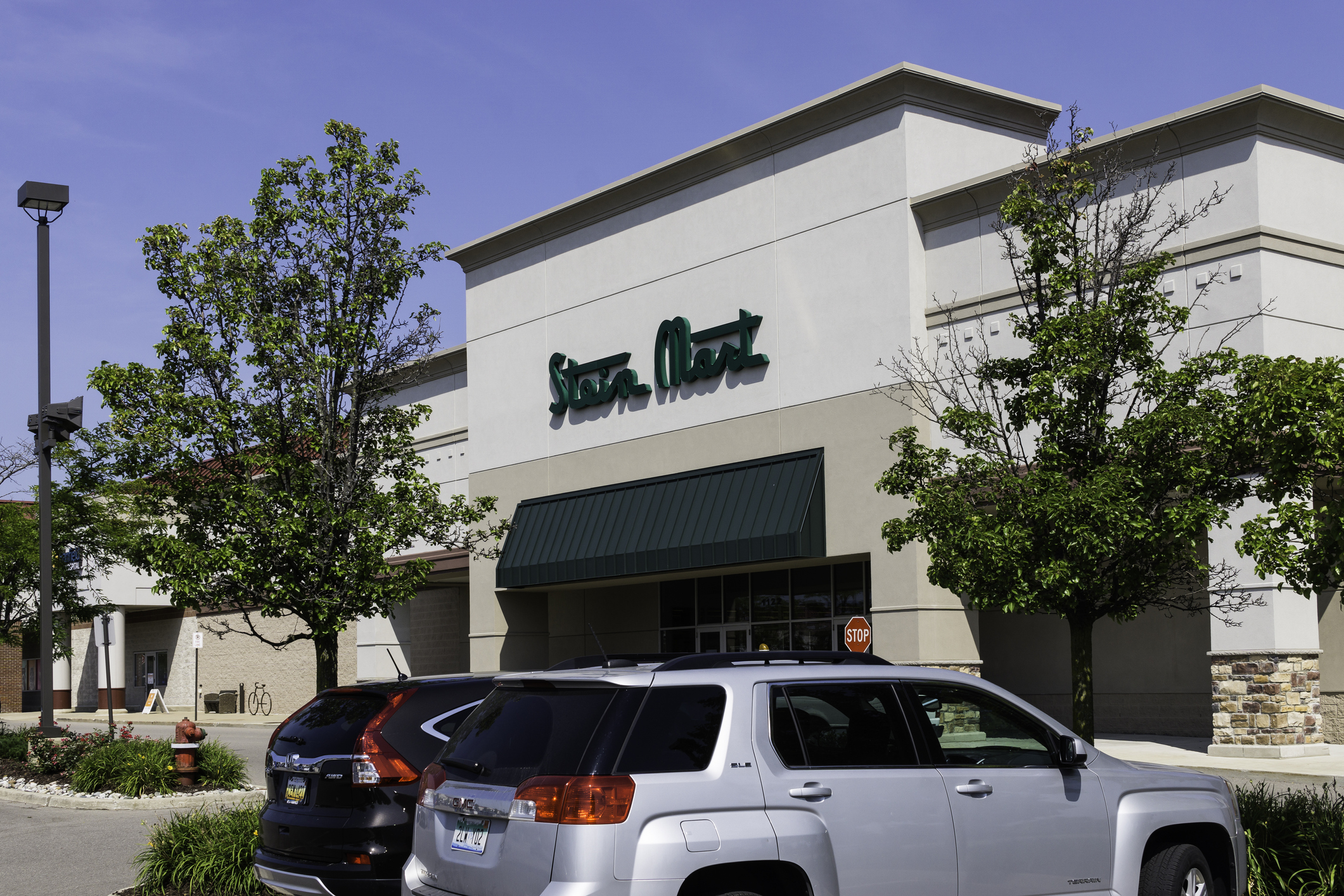 Front exterior of Stein Mart, Rochester Hills, Michigan, a line of cars parked in the parking lot, in a strip mall, another store to the left, on a sunny day, against a blue sky