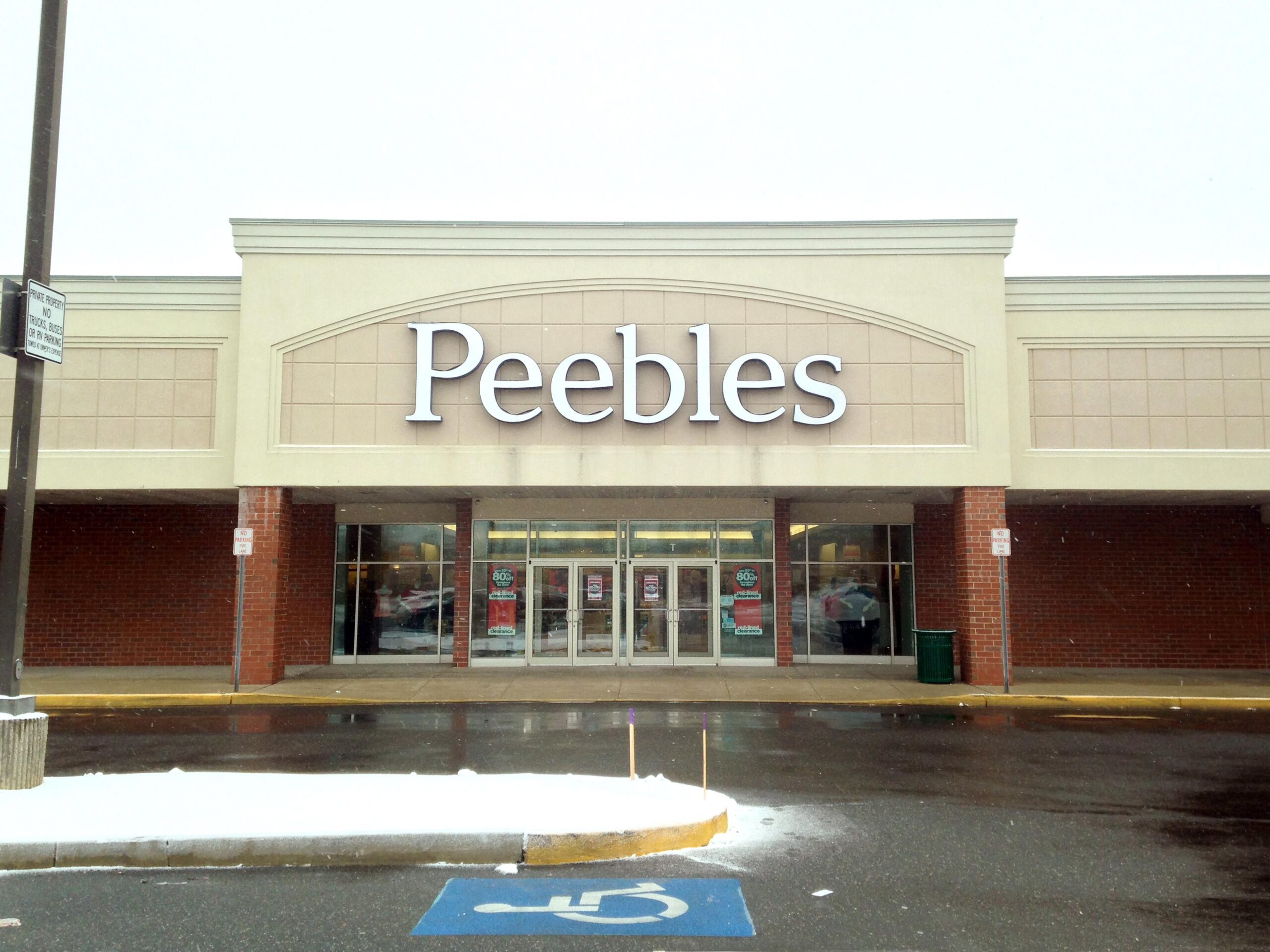 Front exterior of a Peebles retail store, empty feeling in image with a drive-through road and one handicapped parking spot in the foreground, during winter, snow in island and a pale blue sky