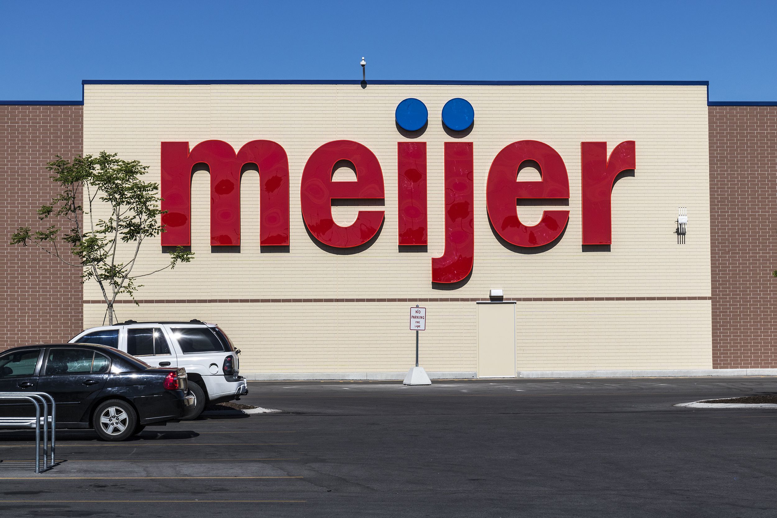 Front exterior of the logo of a Meijer grocery store, Indianapolis, parking lot in the foreground, on a sunny day with a clear blue sky