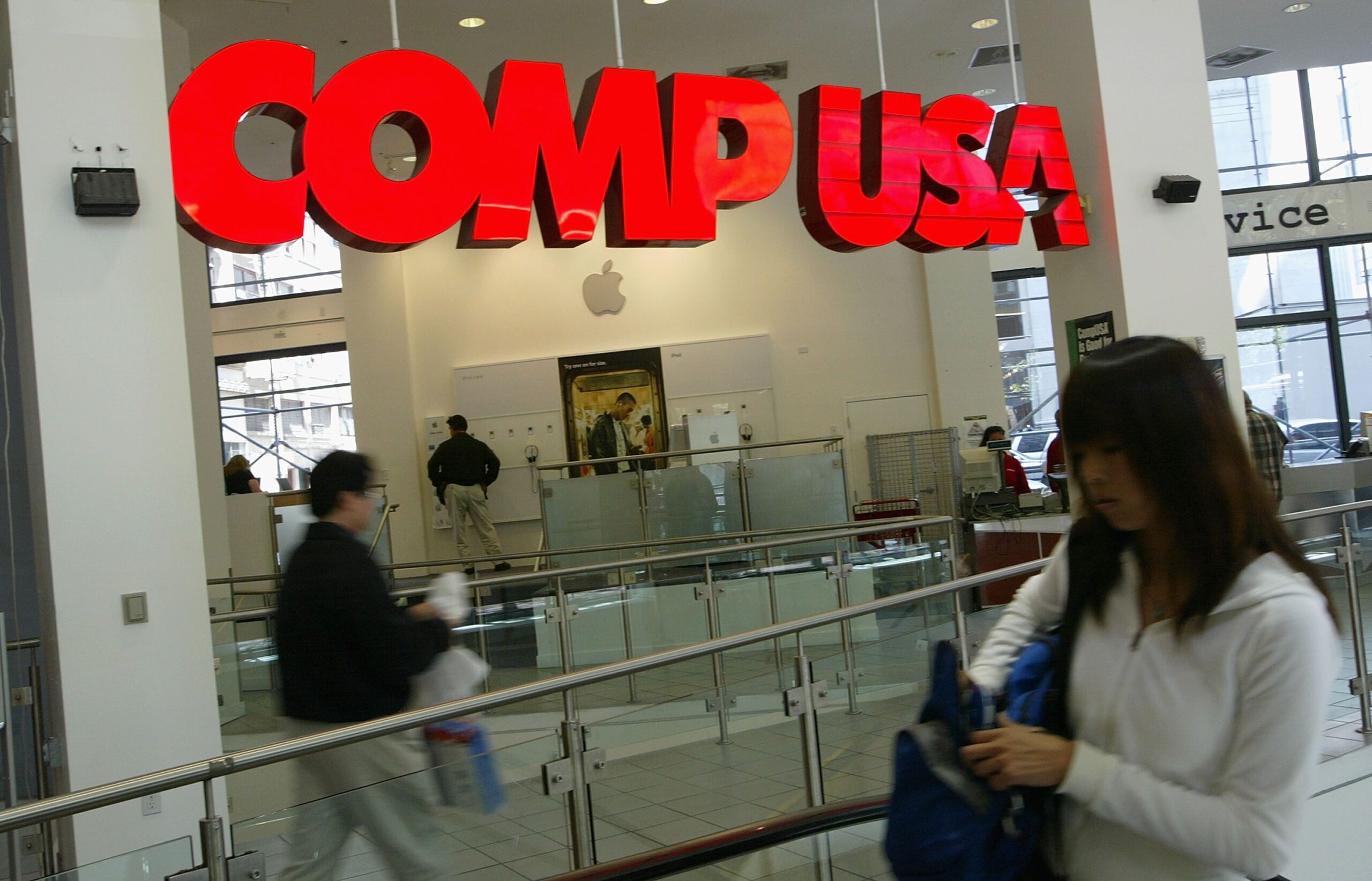 Interior of CompUSA, San Francisco, large red sign hanging, people walking in the foreground, Apple and service sections in the background, 2004