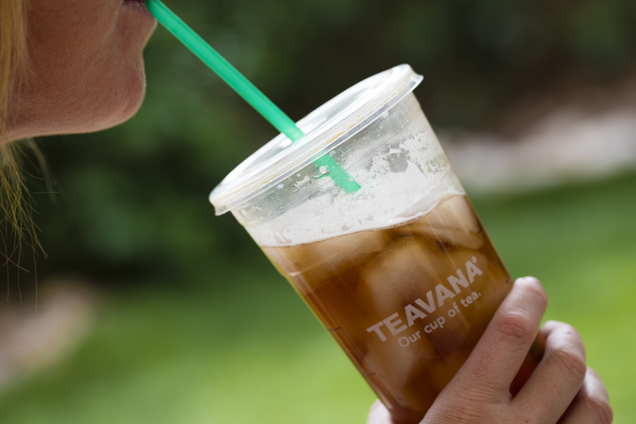 Large Teavana plastic filled with tea, held by a woman drinking it through a green straw, side-view, with a blurred background of grass and trees outdoors