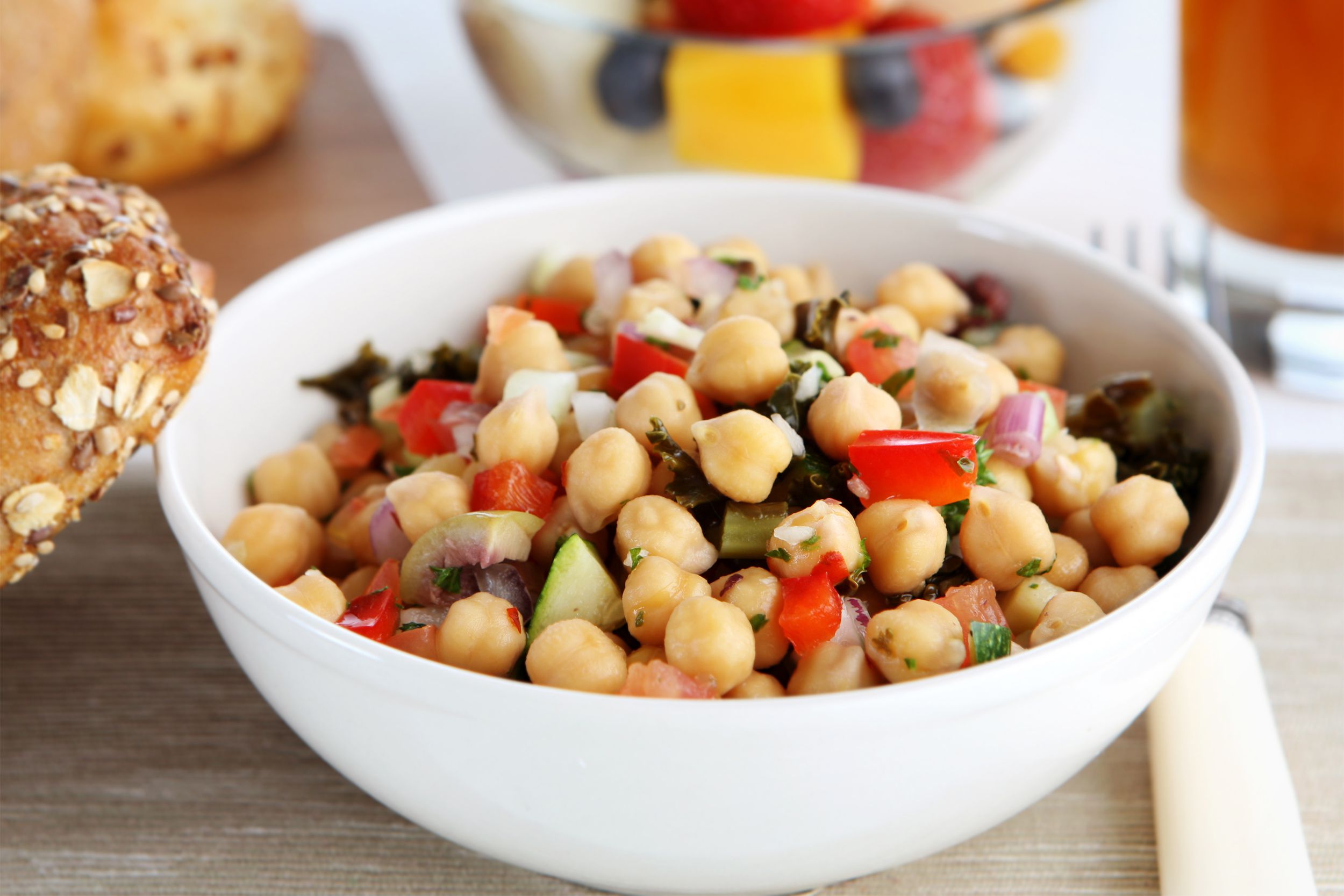 Mediterranean chickpea salad in a white bowl with a small whole wheat bread roll on the left side with a blurred background of whole wheat bread rolls, a fruit salad, and a drink