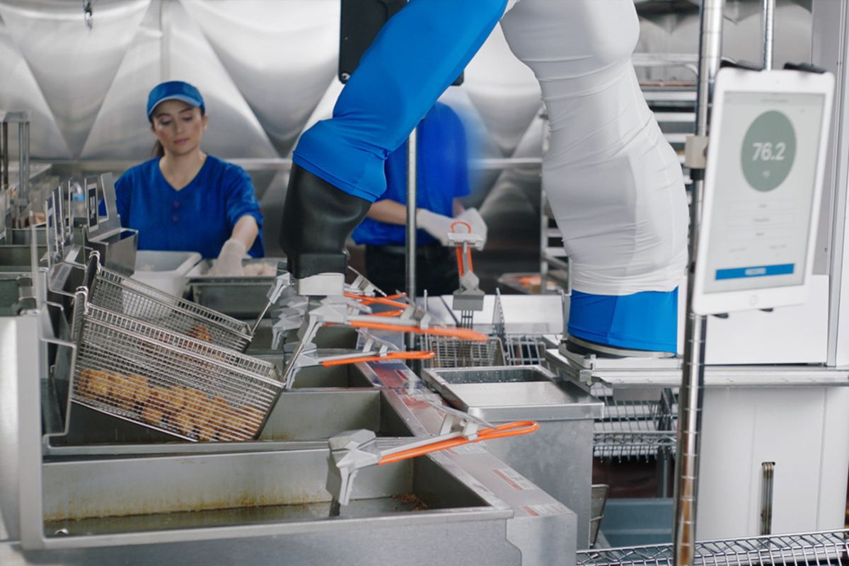Miso Robotics' Flippy Machine at a White Castle kitchen, putting the tater tots fryer in oil, two people working in the background