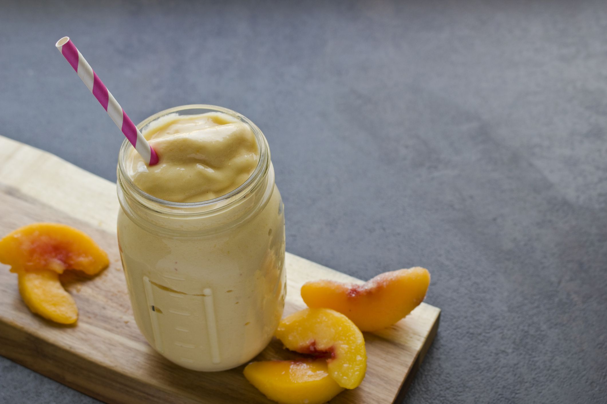 Peaches and cream smoothie in a glass mason jar on a rectangle wooden cutting board with slices of peaches on a granite table