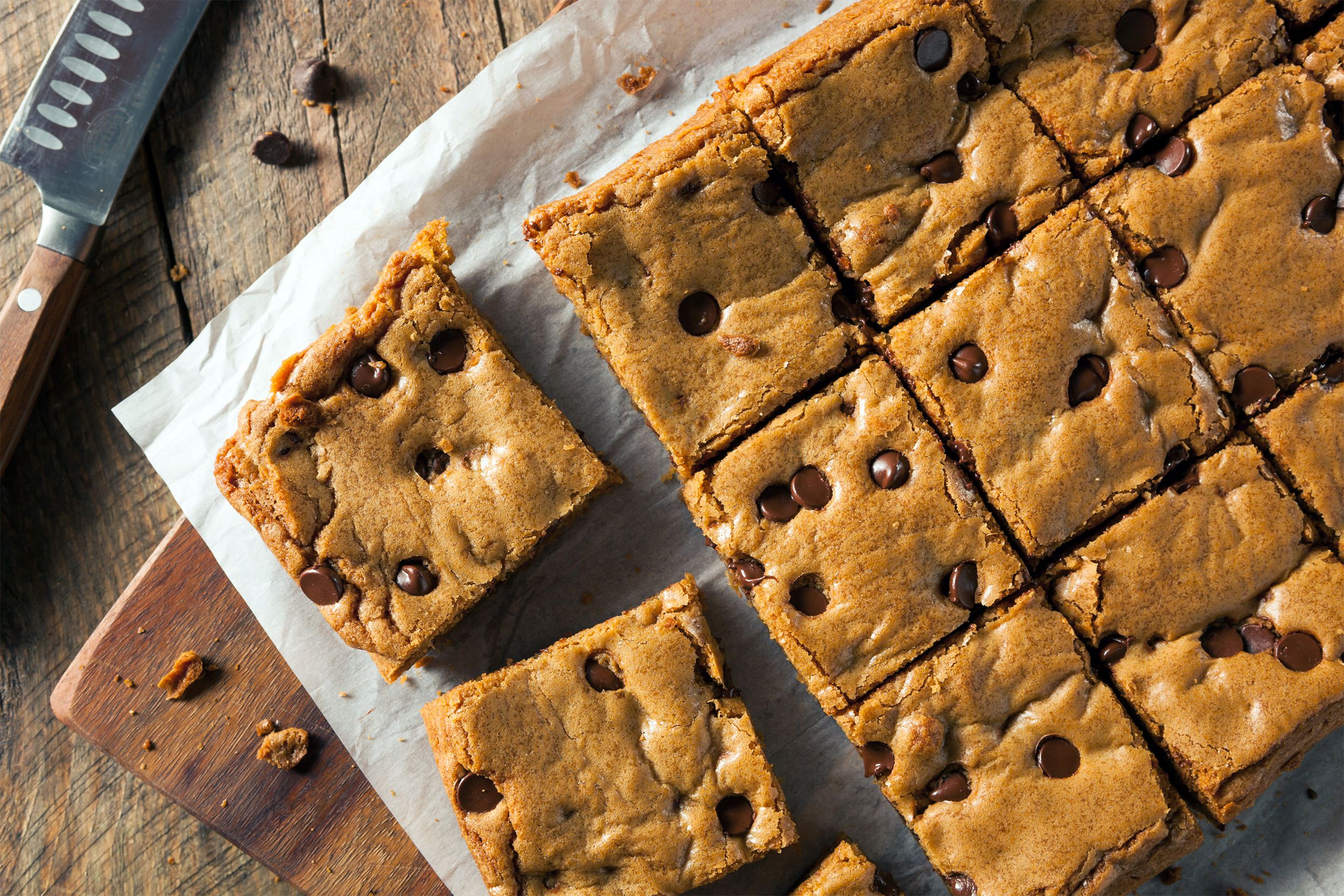 A sheet of Nutella blondies cut into pieces on a white piece of wax paper on a wooden cutting board with a cutting knife on a rustic wooden table