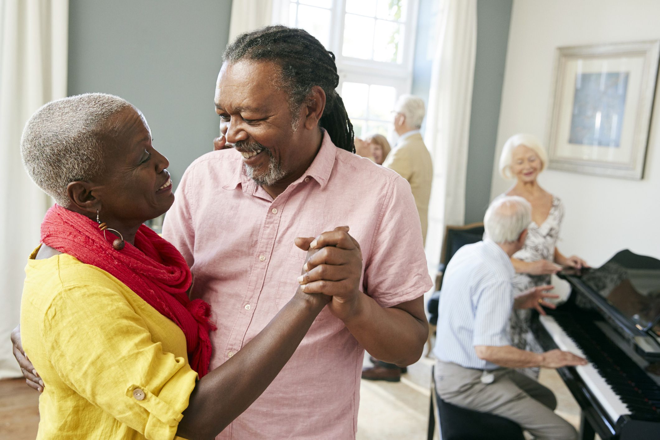 Three senior couples, one in foreground is dancing, one in background man is playing piano with woman, one in background looking out of window