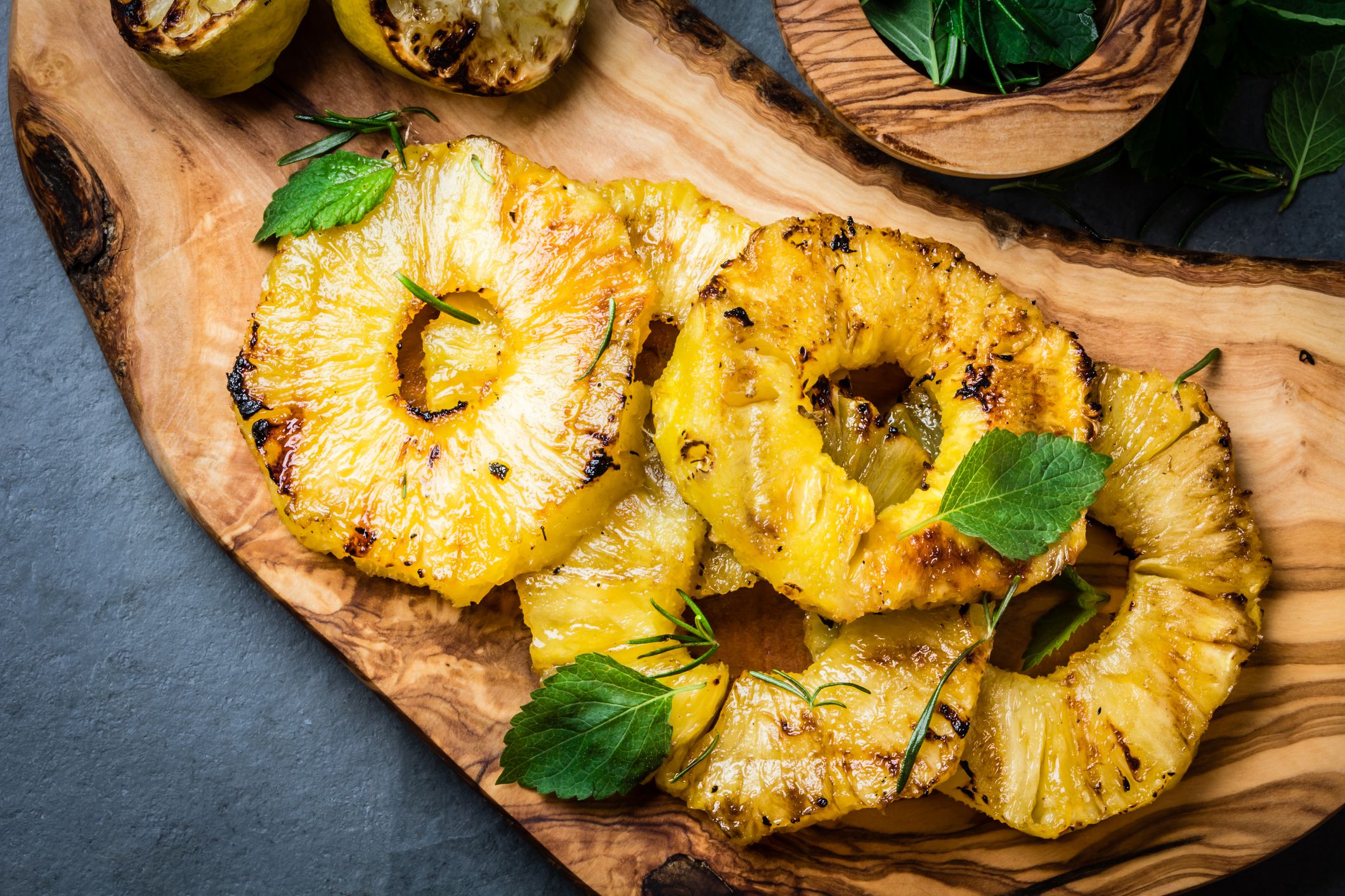 Top view of four grilled pineapple slices on a rustic olive wood board with two garlic slices and an olive wood bowl of herbs on a granite table